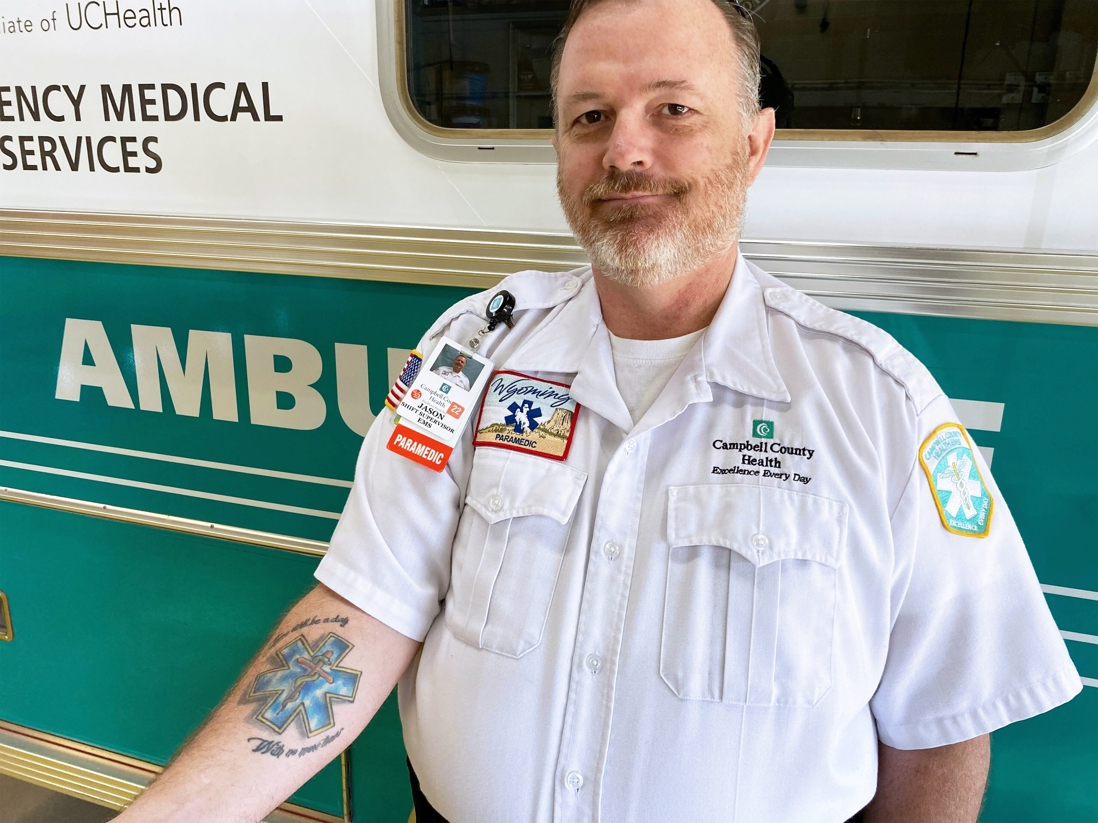 A photo of a male paramedic showing a tattoo on his forearm of the emergency medicine symbol while he poses in front of an ambulance.