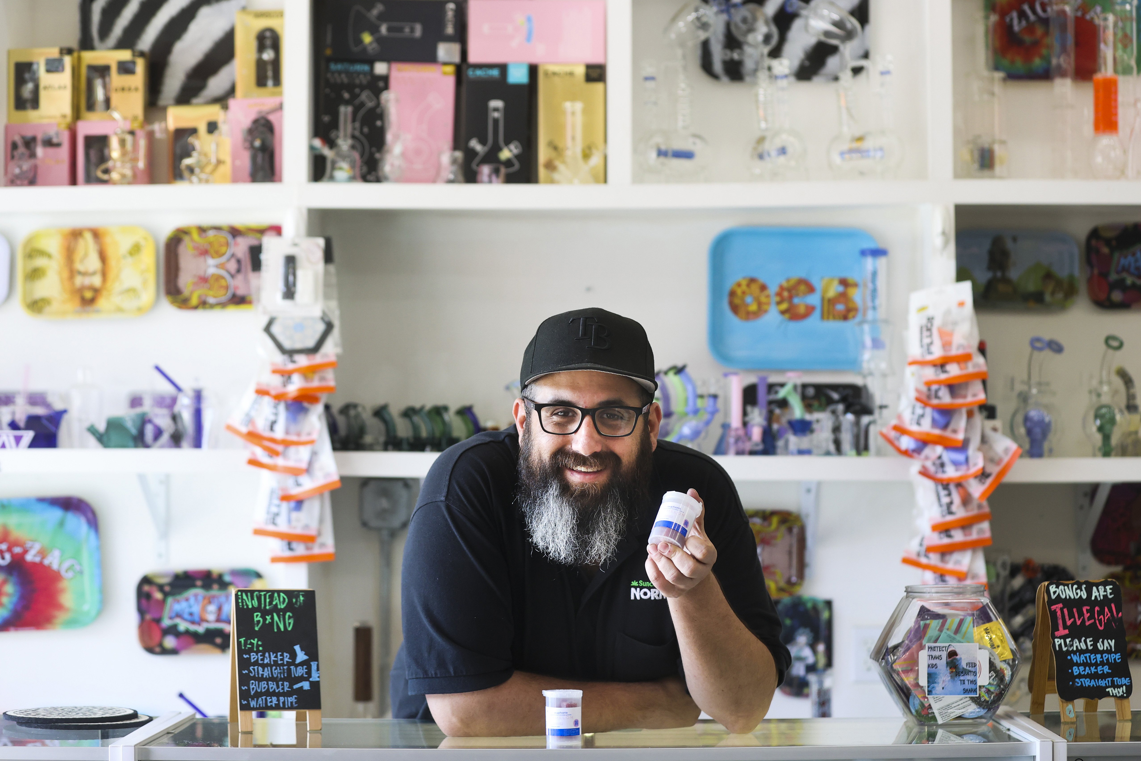 Carlos Hermida, an adult male who wears a baseball cap, black framed glasses, and a beard, smiles kindly as he looks directly at the camera. He is standing behind the counter at his store, and holds a container of Amanita muscaria mushroom gummies in his left hand.