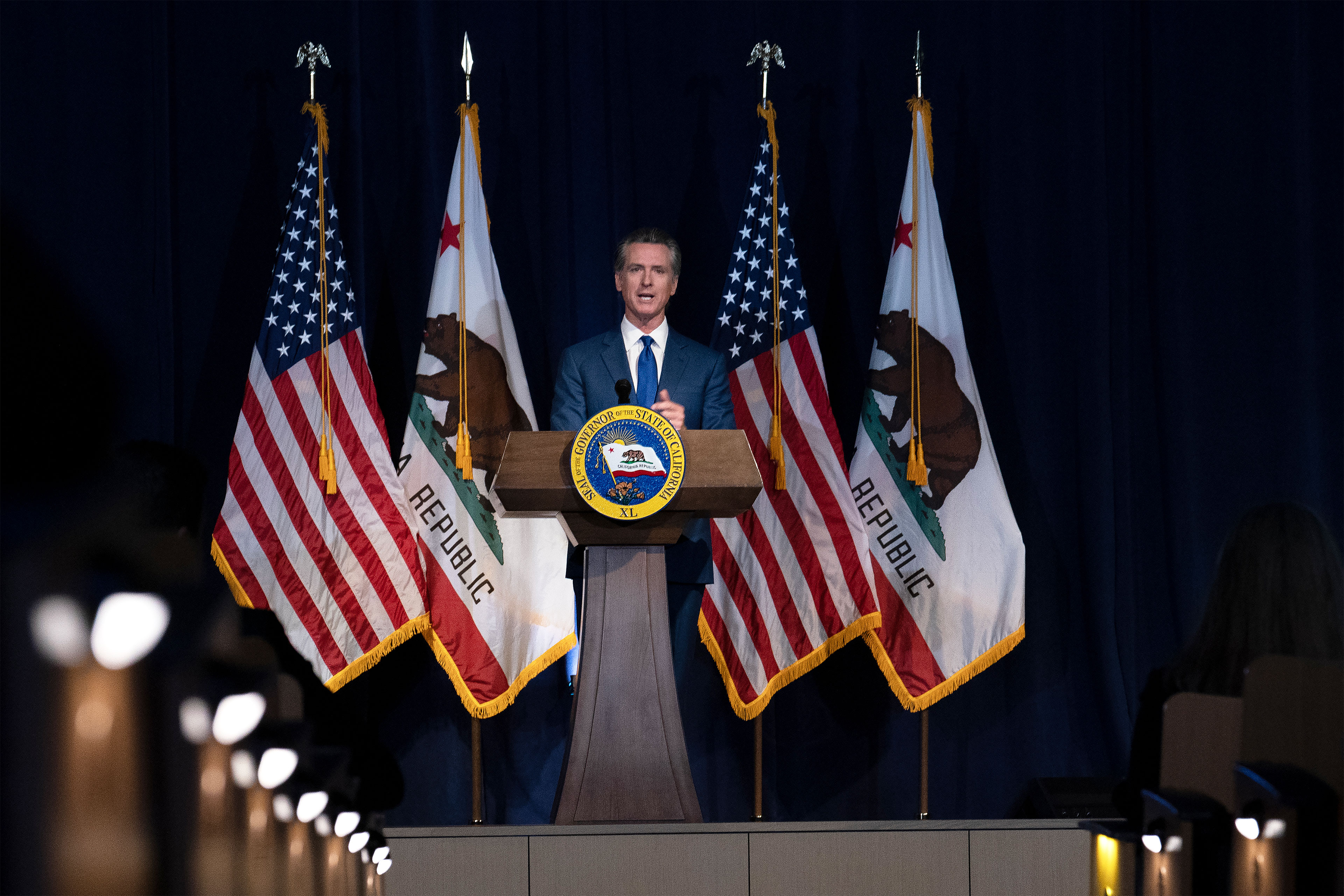 A photo of Gavin Newsom presenting his revised budget behind a podium and surrounded by American and Californian flags.