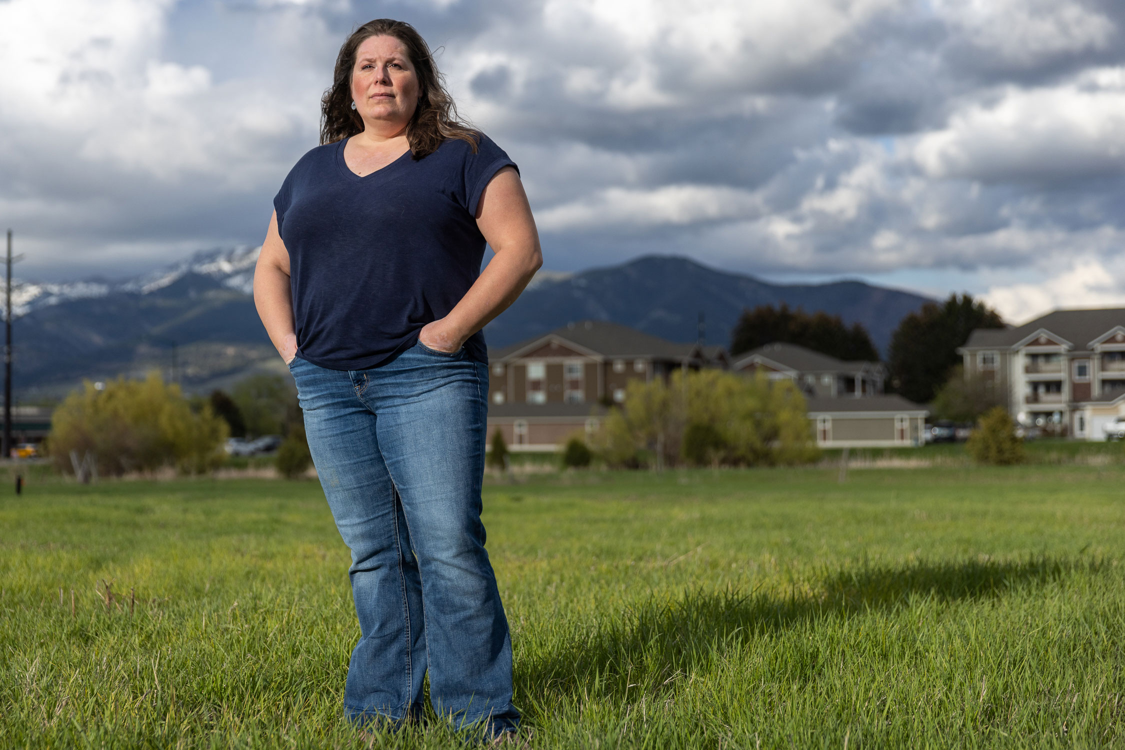 A woman in a dark blue t-shirt and jeans stands in a grassy field. There are buildings beyond the field and mountains farther in the distance.