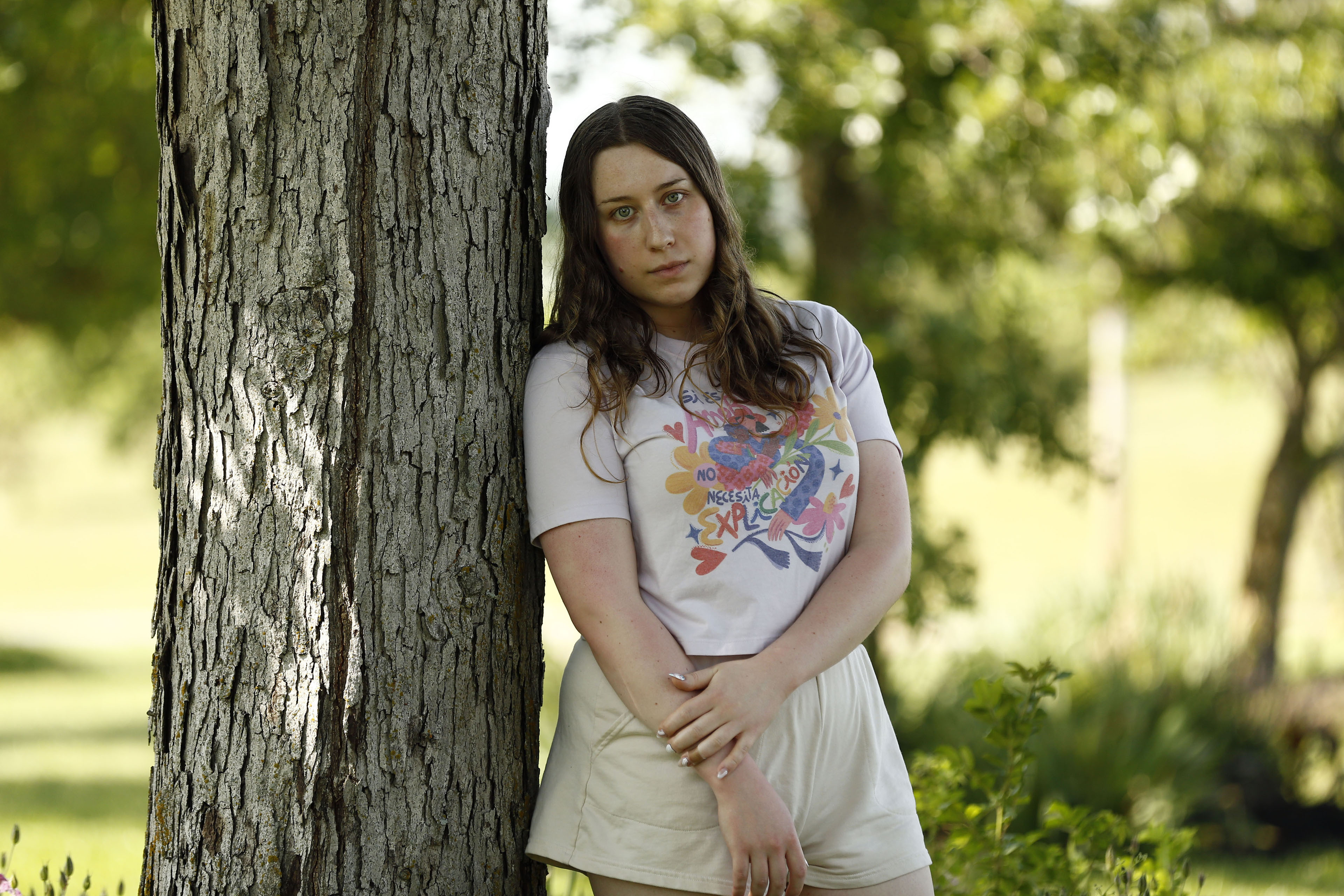 Gwen Schwarz is photographed standing outside on a sunny day. She leans against a tree and looks directly into the camera. She wears a colorful shirt and tan shorts.