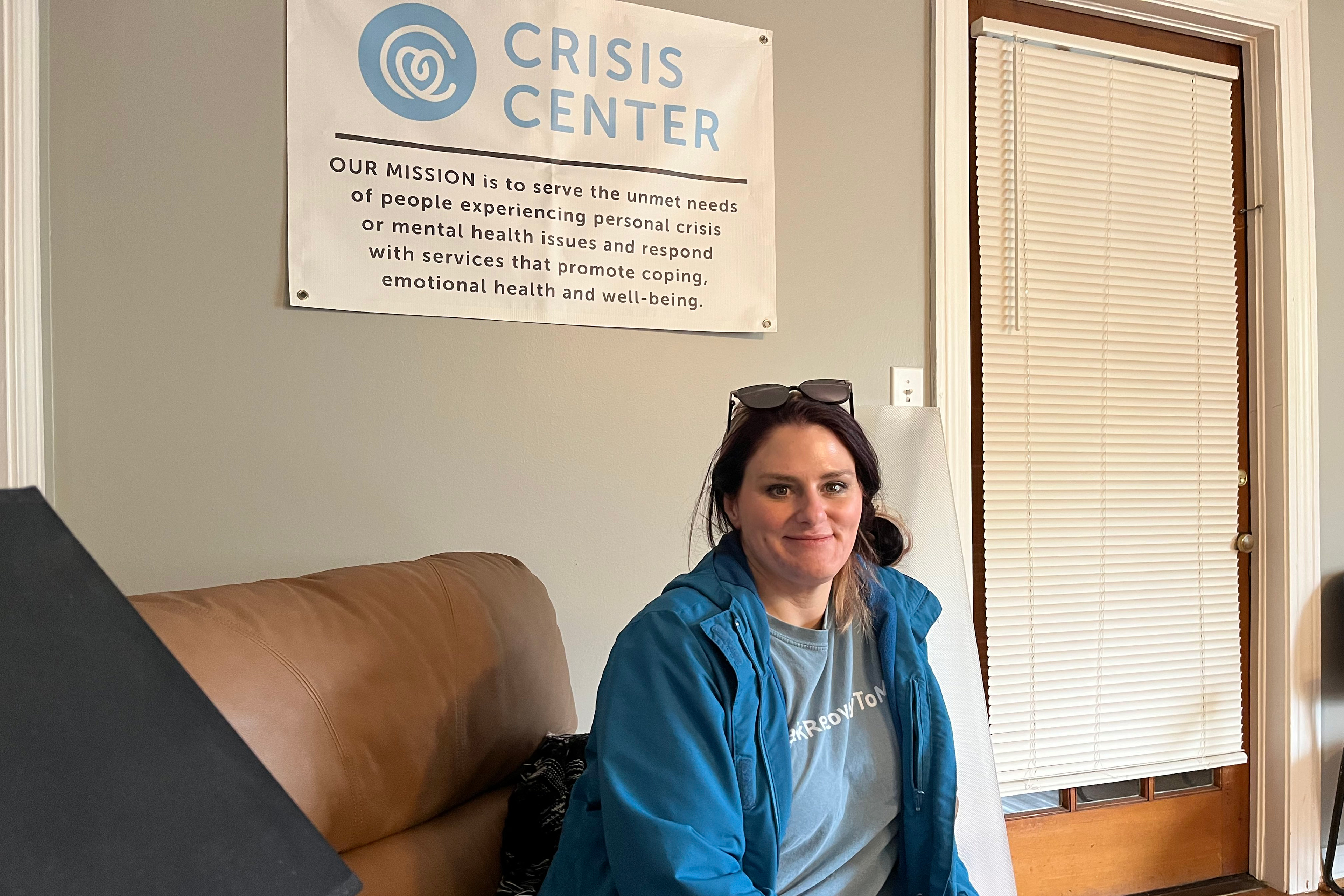 A photo of a woman sitting indoors in front of a banner that reads, "Crisis center."