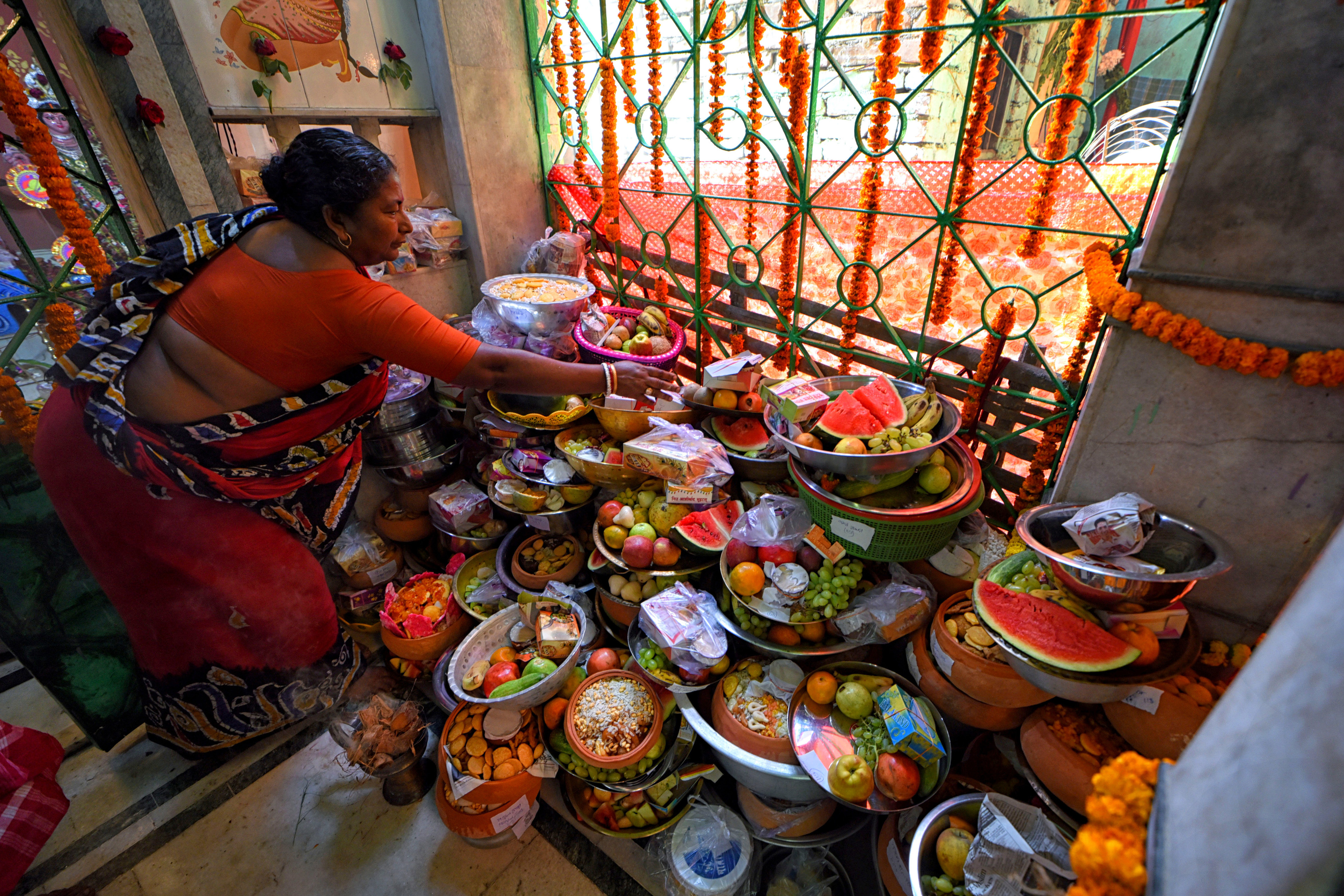 In this vivid, brightly colored photograph, an Indian woman wearing a bright orange sari sets out offerings of food, flowers, and decorative items at a shrine for Shitala Mata. Marigolds are hung in chains around the offerings, and a large window illuminates the room in natural light.