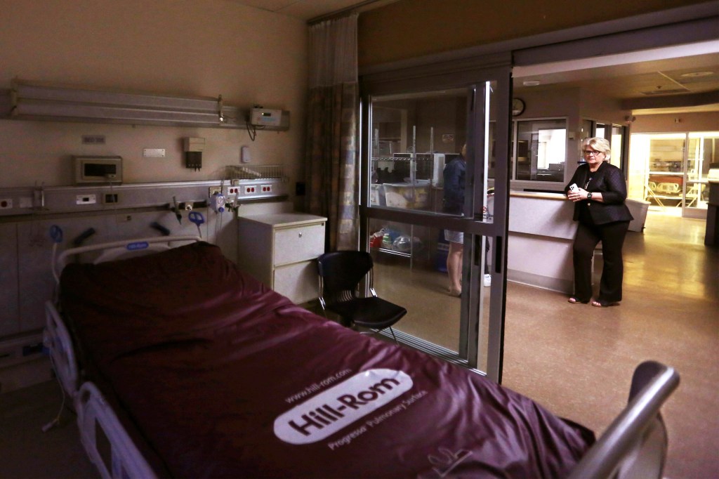 A photo of a woman standing in a vacant hospital as seen through an empty ICU room.