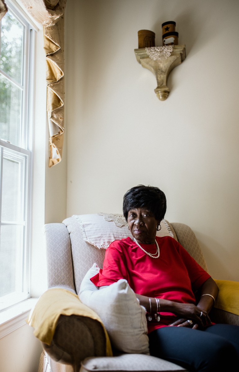 Bluizer Greene sits in a large, plush chair in a sun-lit corner of her home. Her hands are folded comfortably on her lap and she looks directly into the camera.