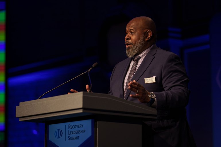 A Black man speaks from behind a lecturn with the words "Recovery Leadership Summit" on the front of it.