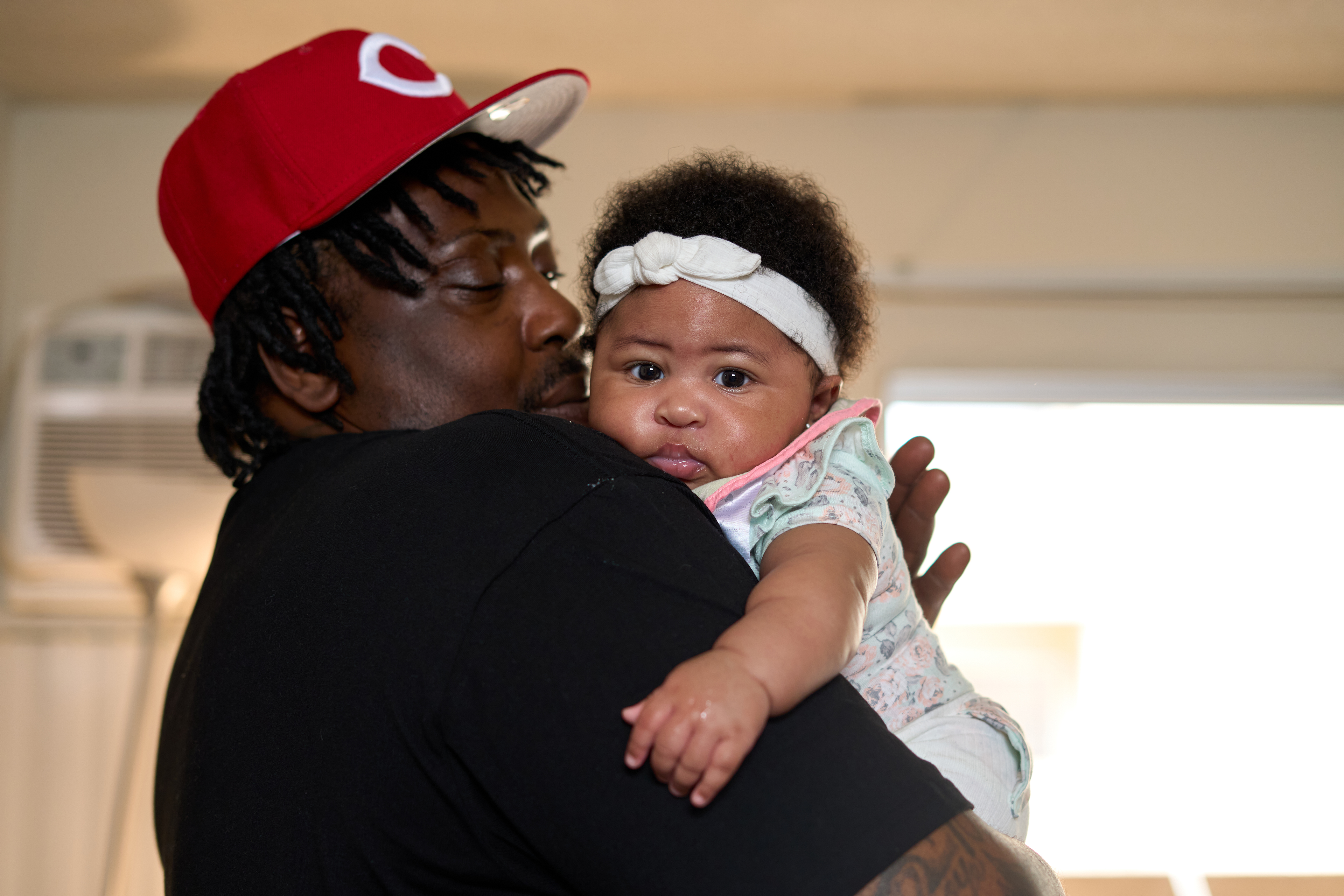A man in a red sports cap, short dreadlocks below, holds a chubby baby across his shoulder. The baby, wearing a white bow headband across her curly dark hair, stares directly at the camera. 