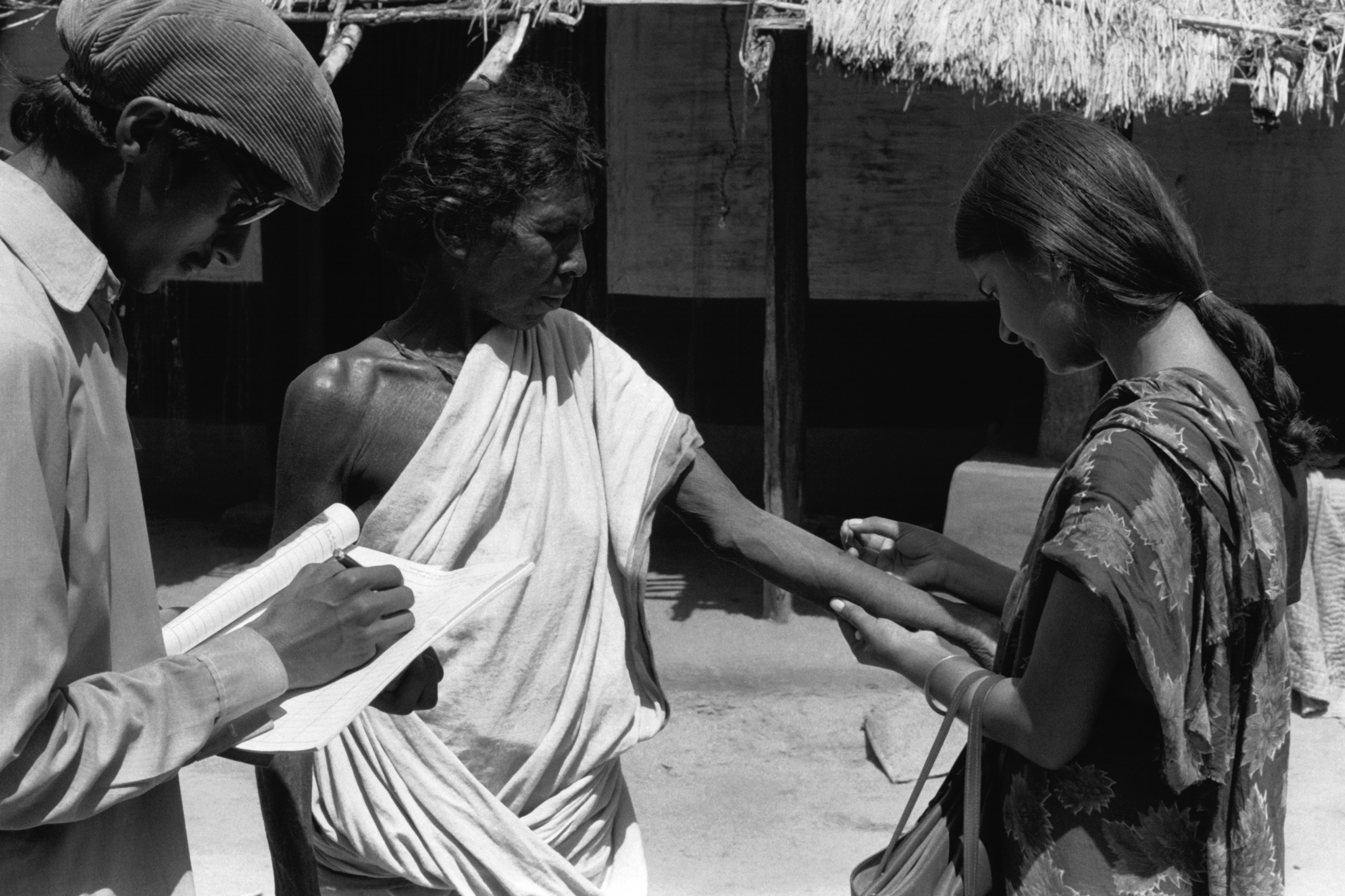 This black-and-white photograph from 1974 shows a woman administering a smallpox vaccine with a bifurcated needle to a man standing outside his home in Bihar, India. Standing beside them is another smallpox containment worker, likely recording the vaccination in a notebook.