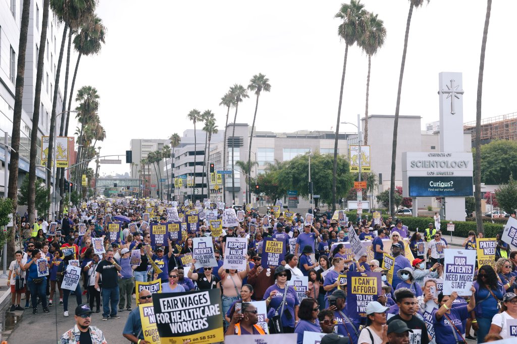 A photo of a crowd of protesters holding signs and marching down a street in Los Angeles. Their signs have slogans such as "Fair wages for healthcare workers," and "Our patients need more staff."