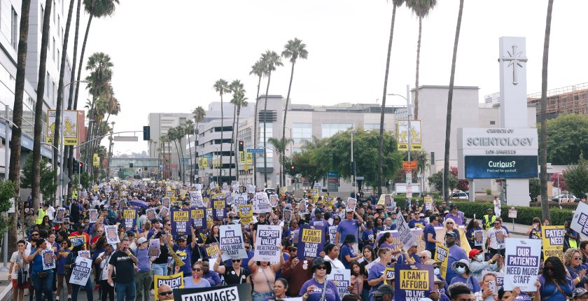 A photo of a crowd of protesters holding signs and marching down a street in Los Angeles. Their signs have slogans such as "Fair wages for healthcare workers," and "Our patients need more staff."