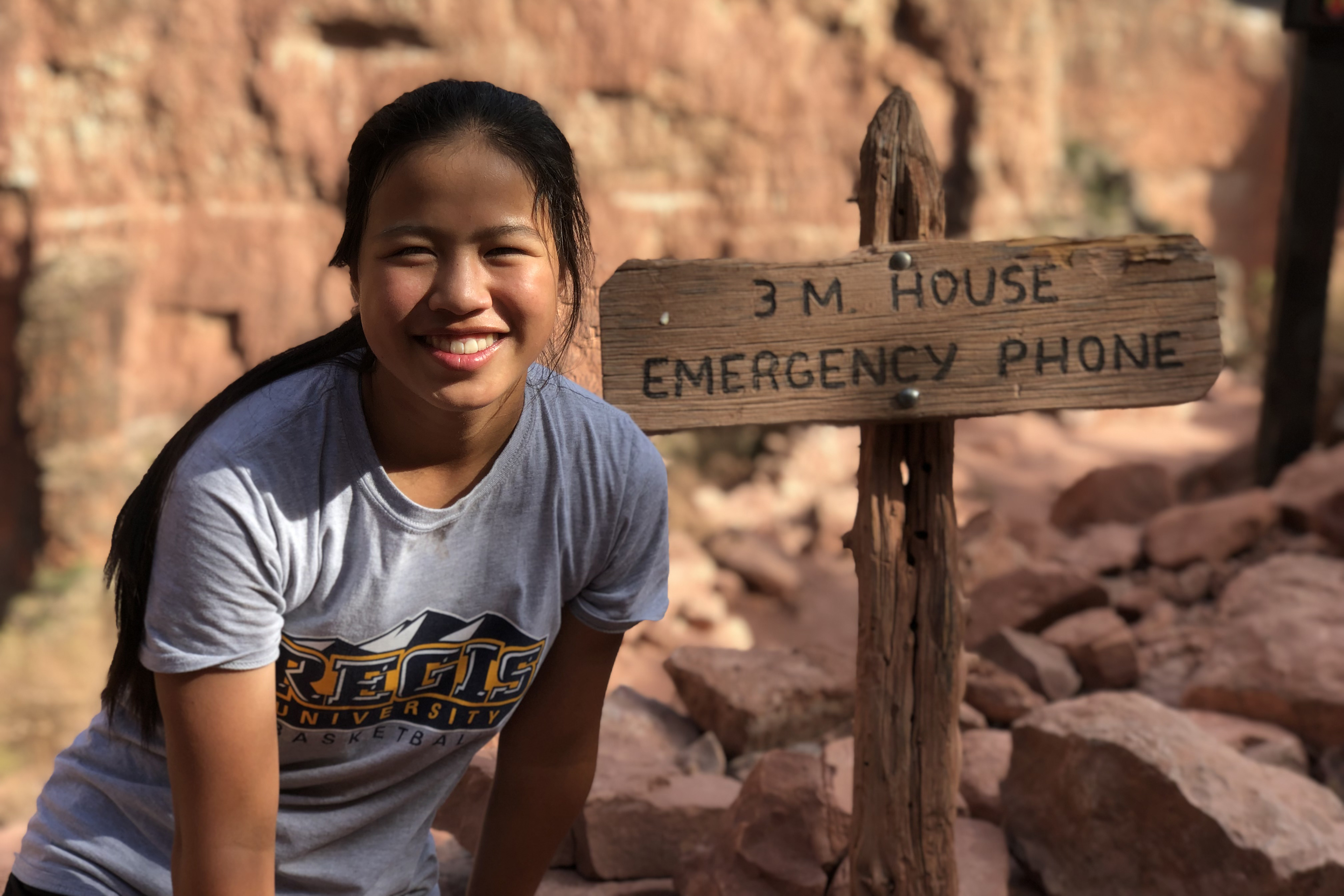 Natalie Lin, a high school student, is sitting outdoors and smiles at the camera. Beside her, a sign reads: "3M House Emergency Phone"