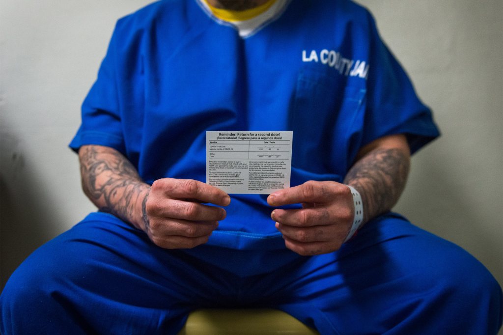 A man in blue prison uniform sits and holds out a card in front of him with both hands.