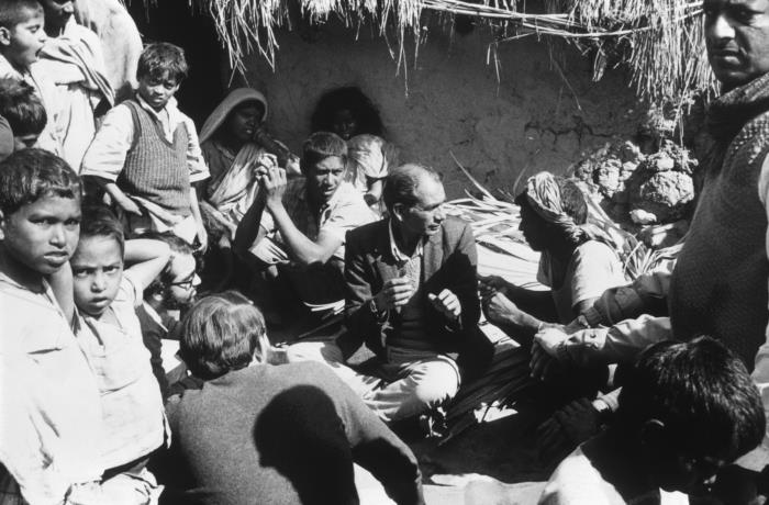 A black-and-white photograph from 1975 shows a health care worker sitting on the ground among a small crowd as he speaks to them.