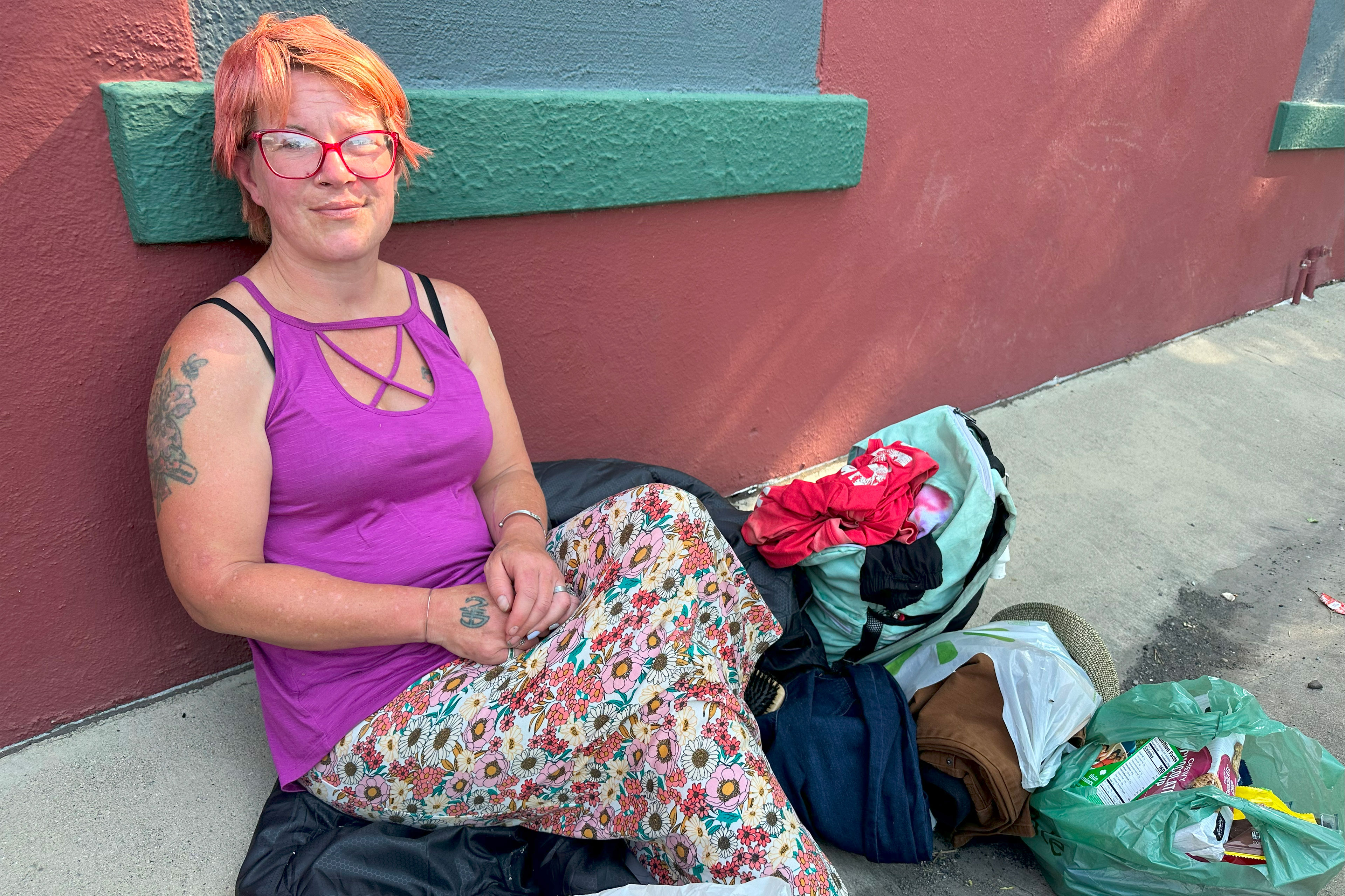 A woman sits on a jacket on a sidewalk and leans against a building. She has short hair and wears red glasses, a purple tank top, and a long skirt with a flower print. There are a few bags beside her.
