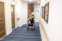 A photo of an elderly woman walking down a hallway with a walker.