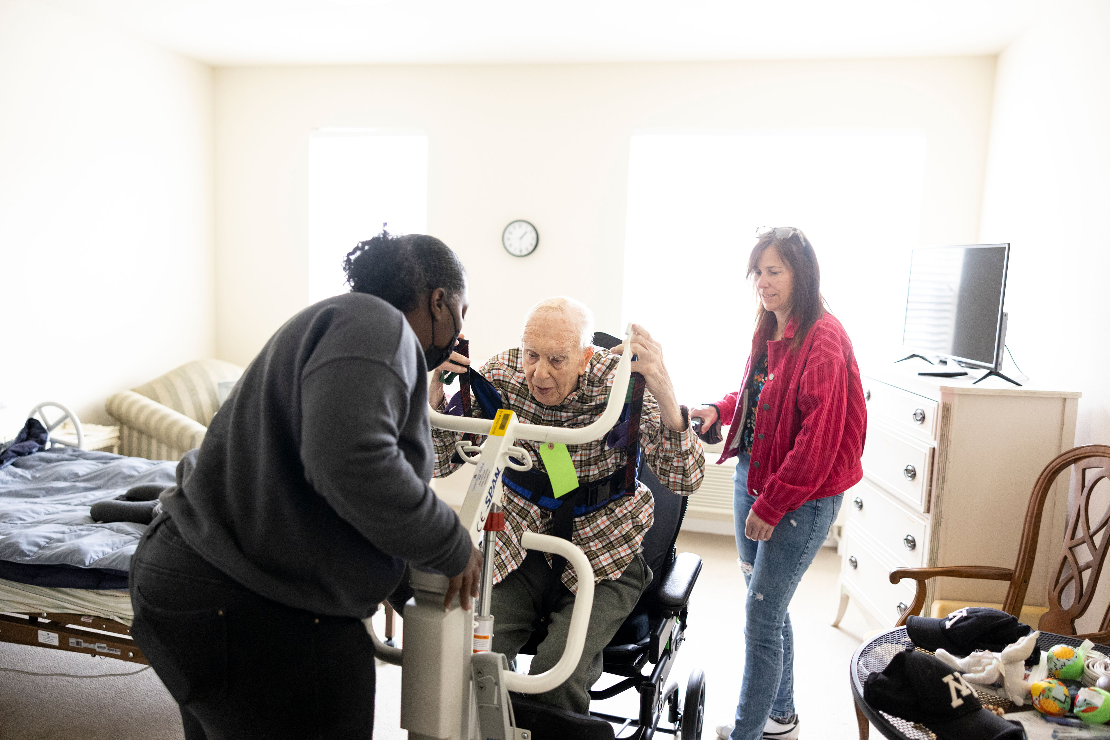 A photo of an elderly man being helped into a wheelchair by a nursing aid. A woman is watching from behind the man.