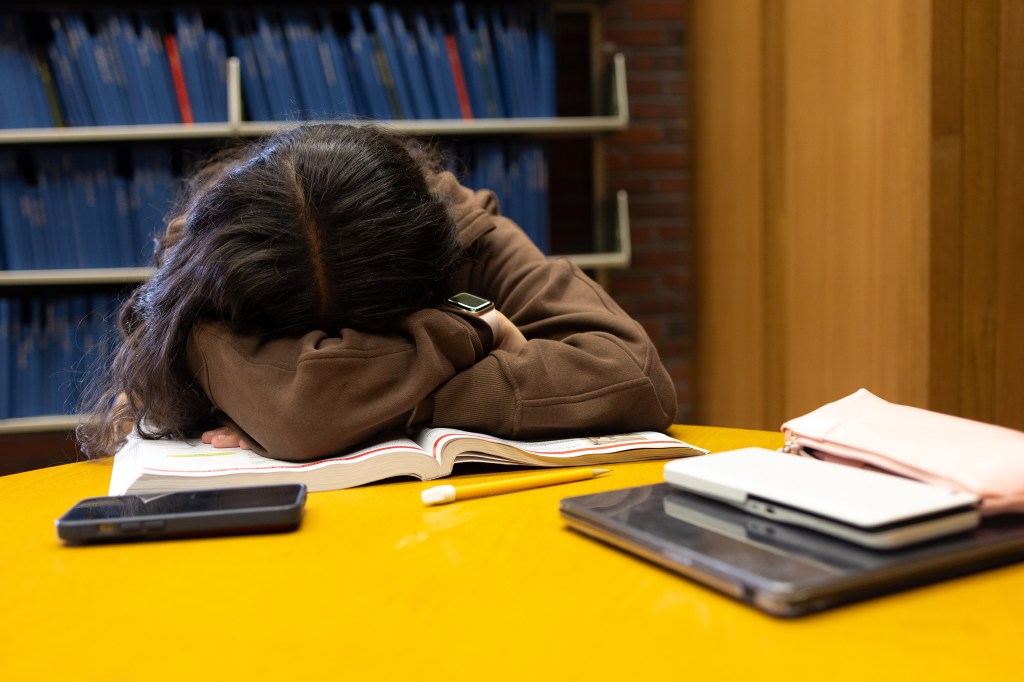A student, who appears to be sleeping, has their face down in her arms as they sits at a desk.