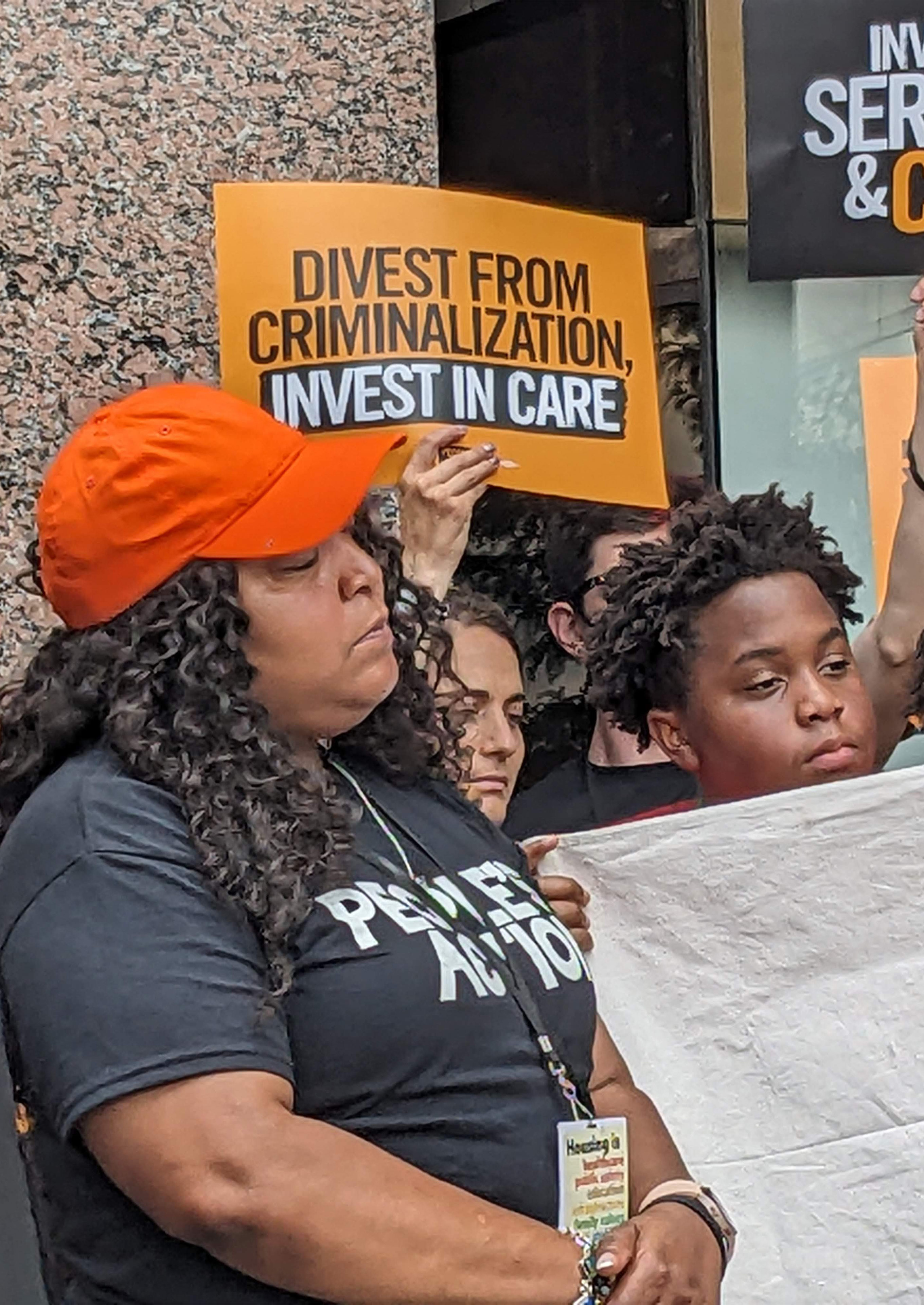 Shameka Parrish-Wright stands amongst others in front of the Drug Enforcement Administration Building. A person behind her is holding up a sign that reads, "DIVEST FROM CRIMINALIZATION, INVEST IN CARE."