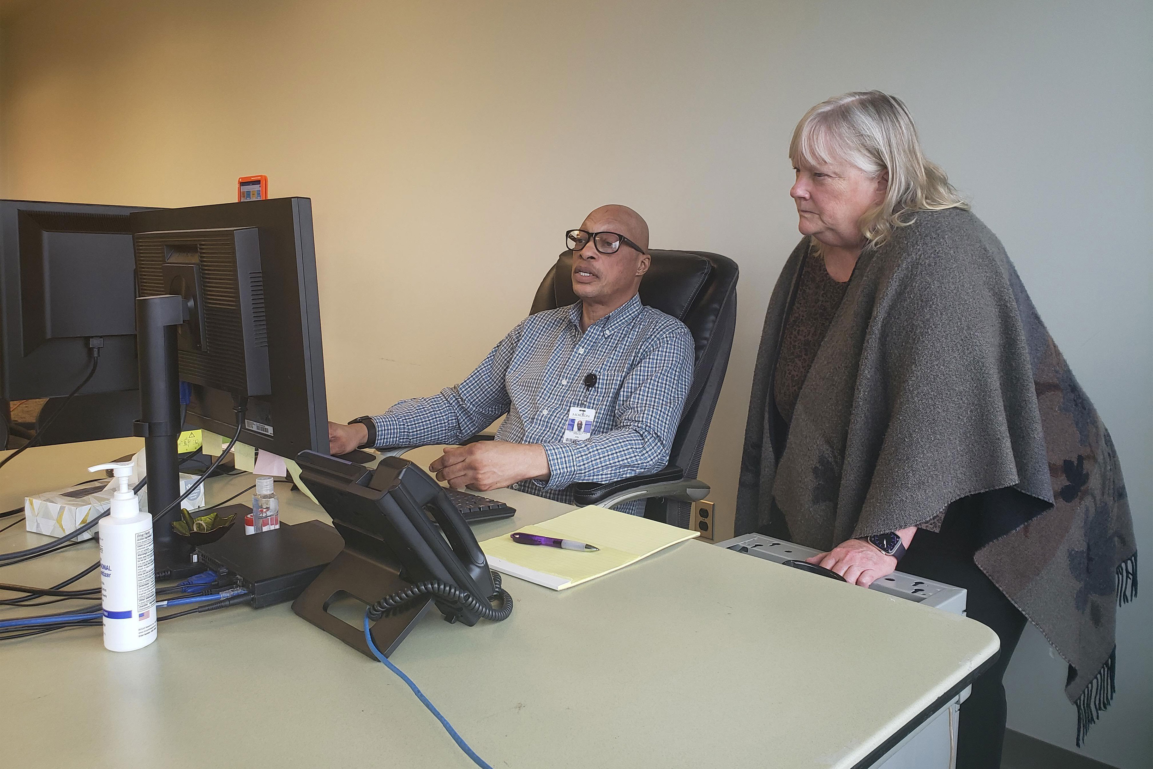 A man sits at a computer on a desk and a woman stands next to him and looks at the computer screen.