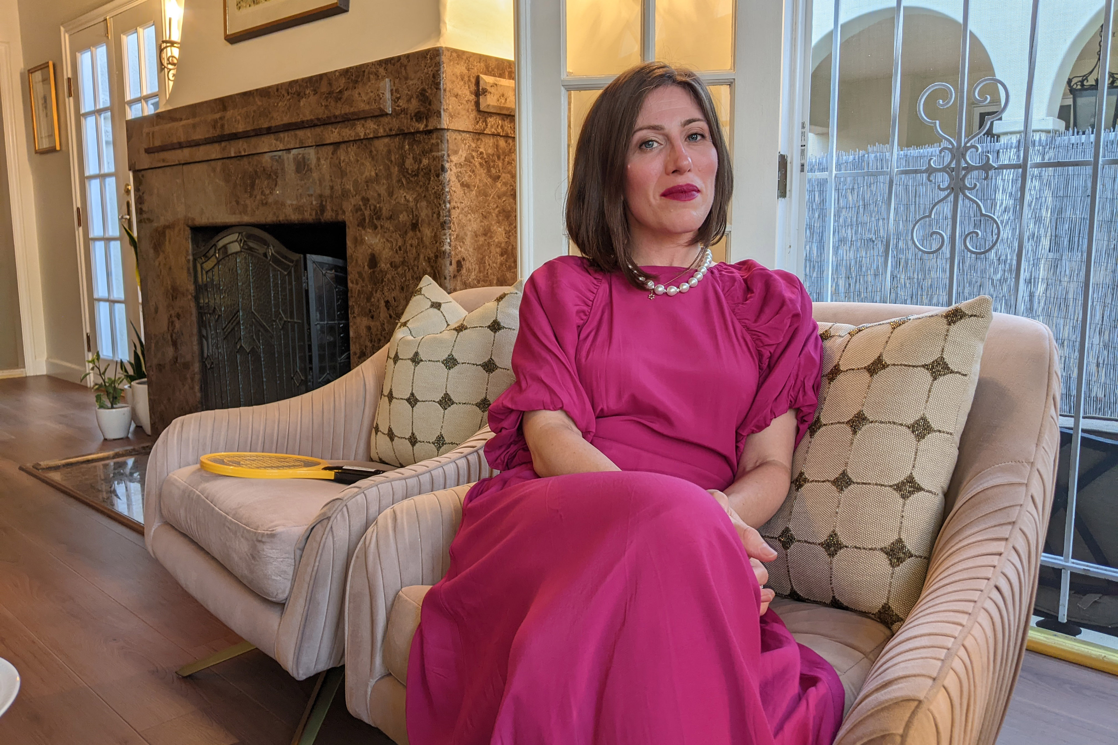 Ariella Morrow, sits on a chair in her home. She wears a bright pink dress and a pearl necklace.