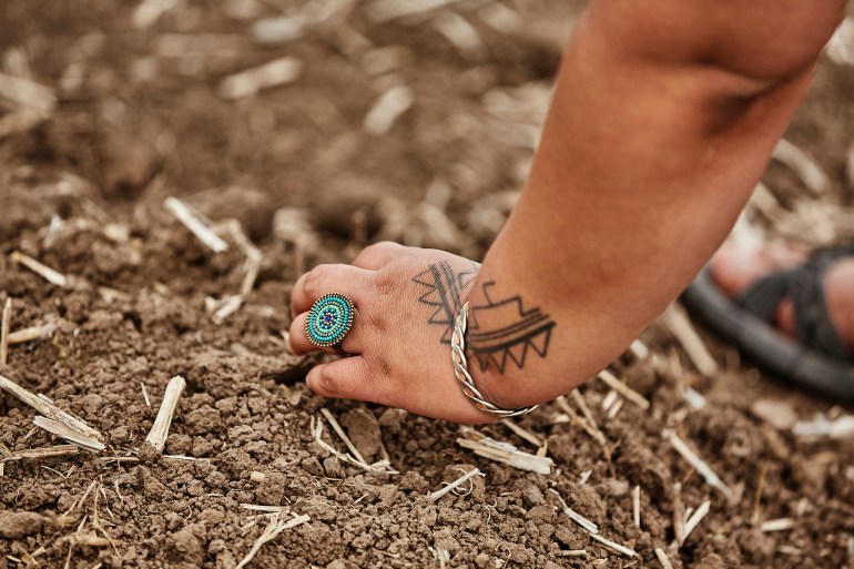The tattooed hand of a woman, wearing a circular turquoise ring and a bracelet, digs in dirt in a garden.