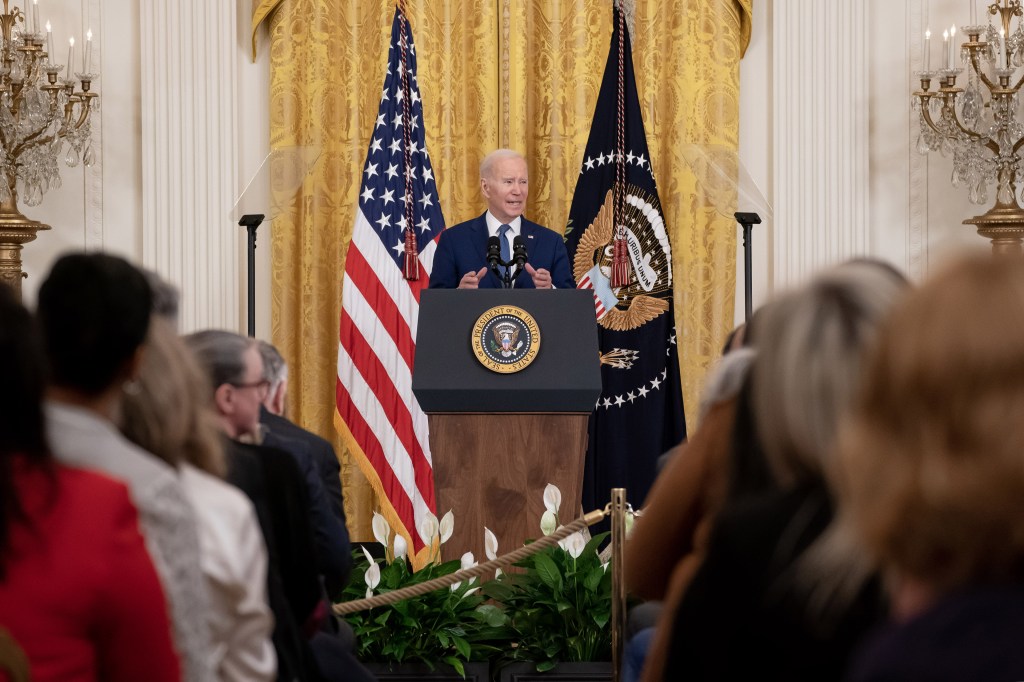 U.S. President Joe Biden is standing at a presidential podium in front of a seated audience in the White House.