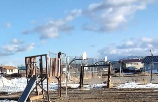 A photograph of an outdoor playground in Heart Butte, Montana. The playground has a fort and slide, as well as basket ball hoops. Snow partially covers the ground.