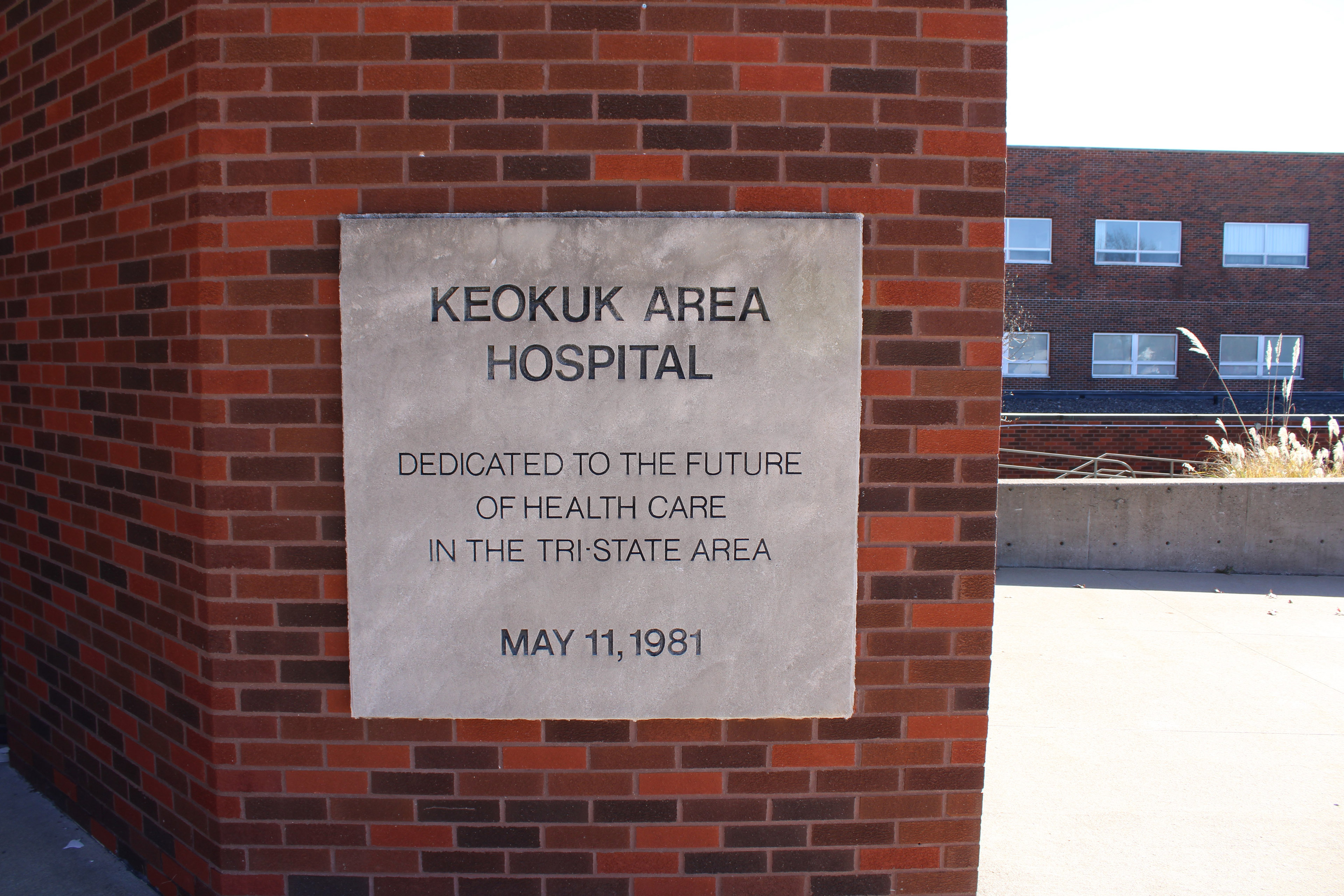 A engraved concrete sign on a brick wall of the Keokuk Area Hospital reads, "Keokuk Area Hospital / Dedicated to the future of health care in the tri-state area / May 11, 1981"
