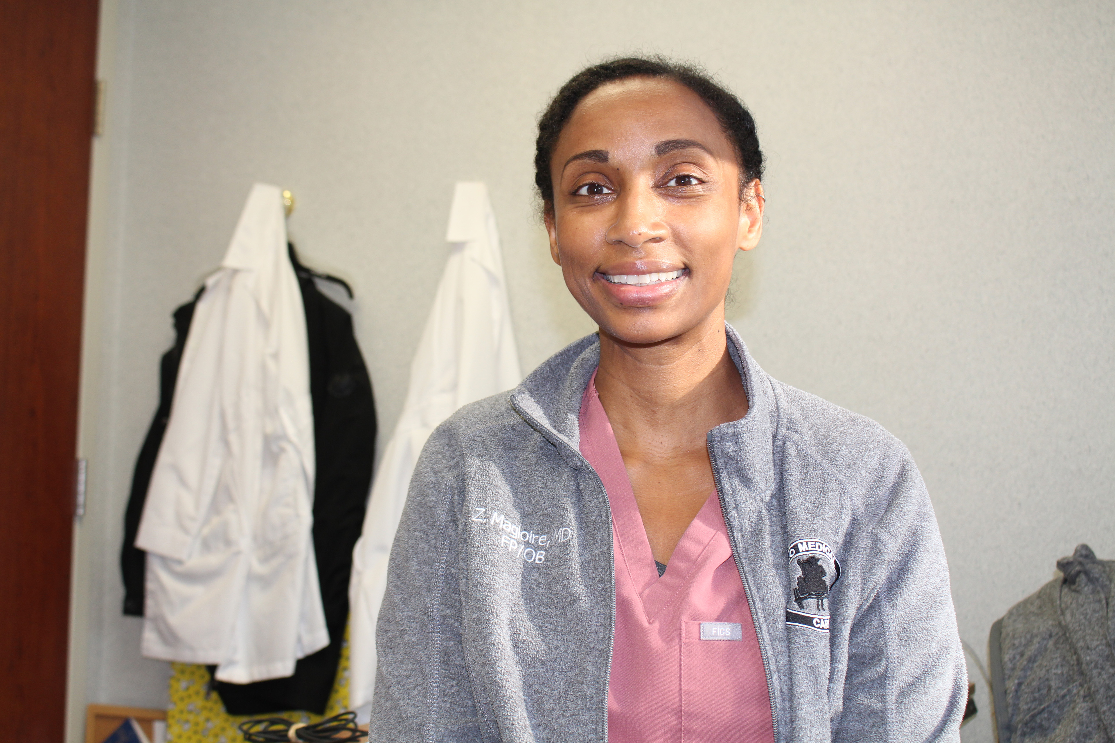 Zita Magloire, wearing hospital scrubs and a fleece jacket, smiles towards the camera.