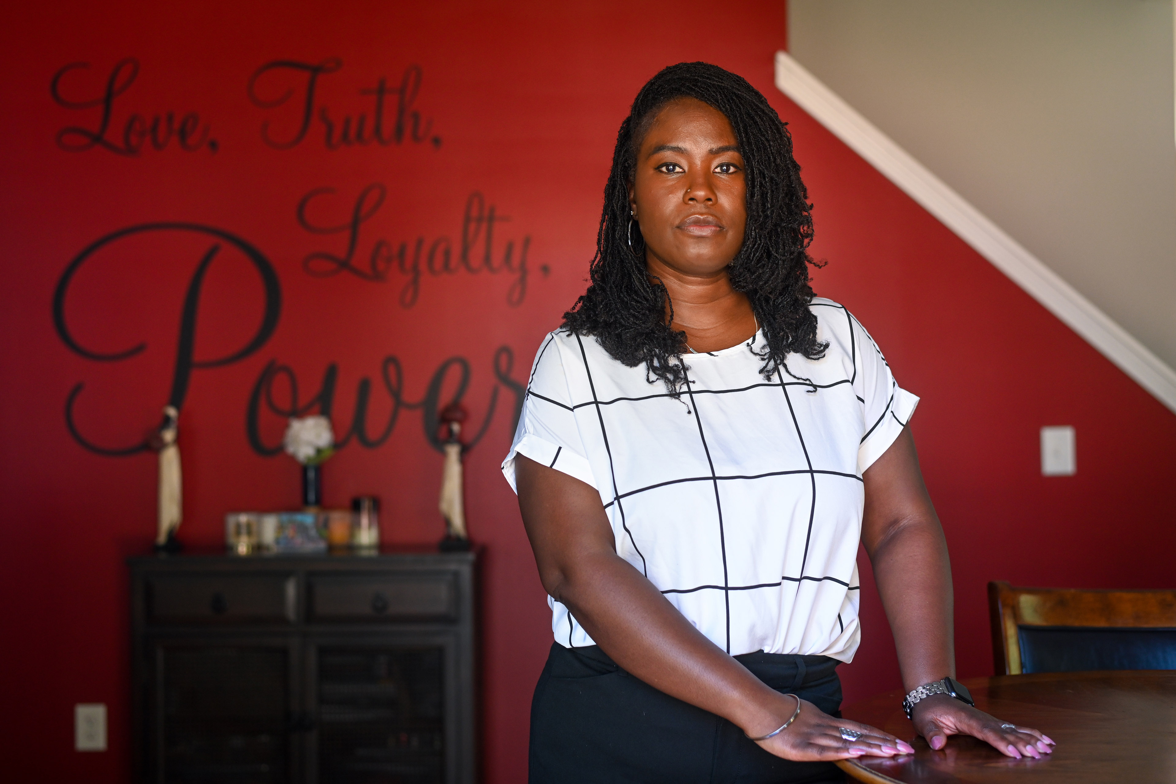 Charity Watkins stands in her home. Behind her on a red wall, large words in elegant script read, "Love, Truth, Loyalty, Power."