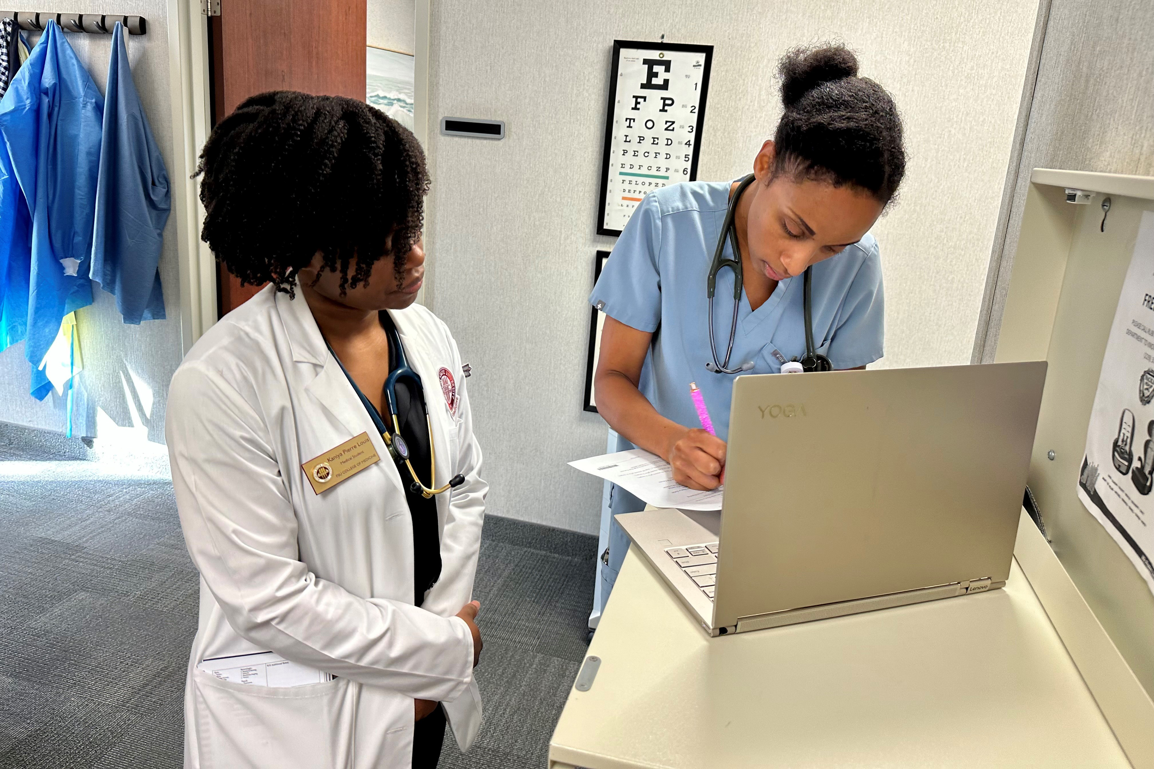 Kaniya Pierre Louis (left) stands beside Zita Magloire (right) as she works at a computer. Both are in a hospital setting and wearing scrubs. Kaniya Pierre Louis wears a white doctor's jacket.