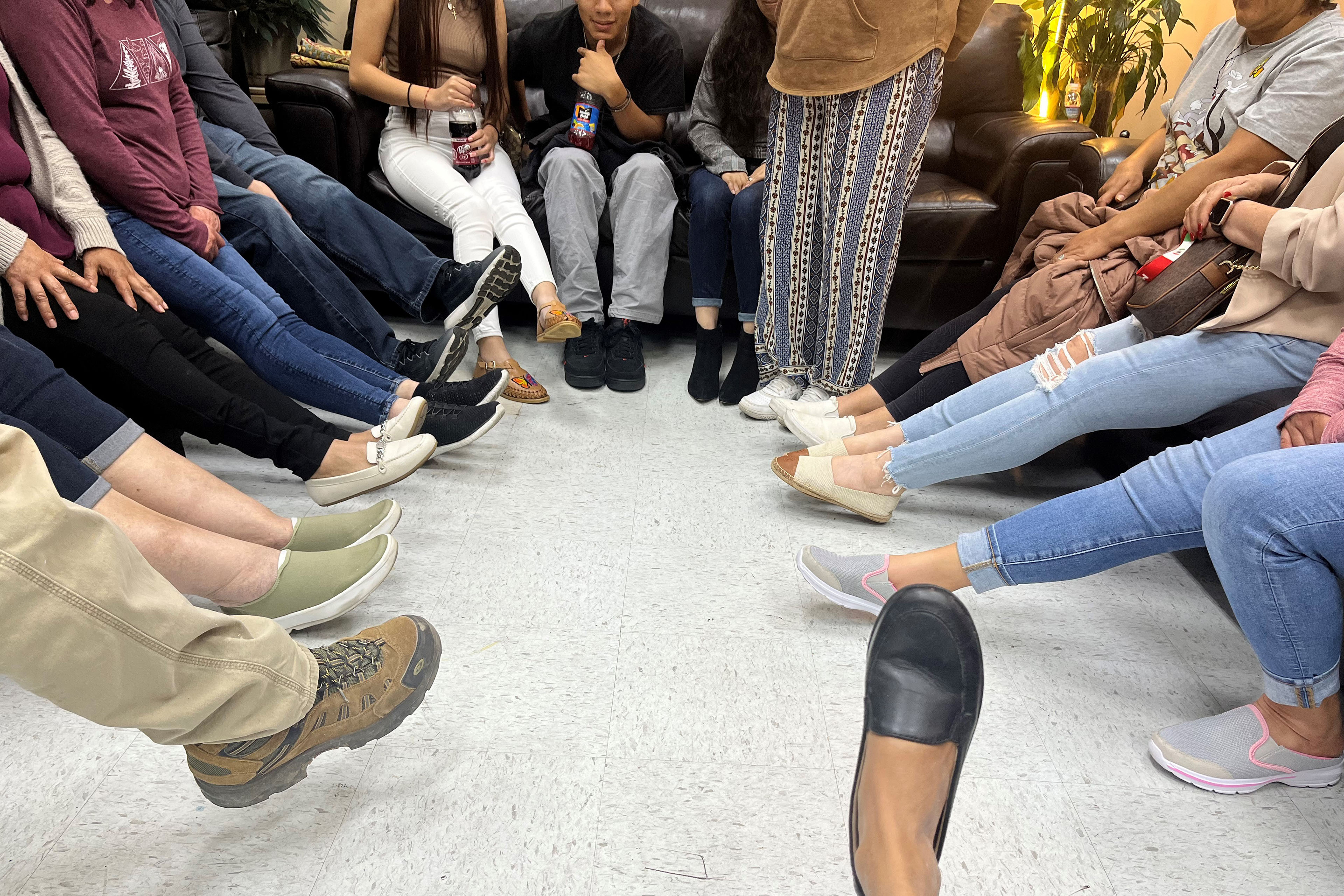 A photograph of a group of people sitting on chairs in a circle from the legs down. The group all stretches out their feet to the center of the circle.