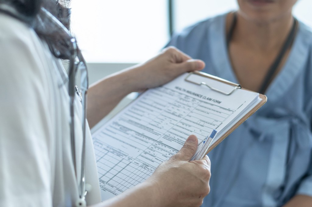 A photo of a doctor holding a clipboard in front of a patient labeled, "Health insurance claim form."