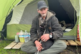 Samuel Buckles, an older man, sits on the ground outside with his dog in his lap. The small black dog reaches up to lick Buckles' face. Behind them is a green tent, where Buckles resides.
