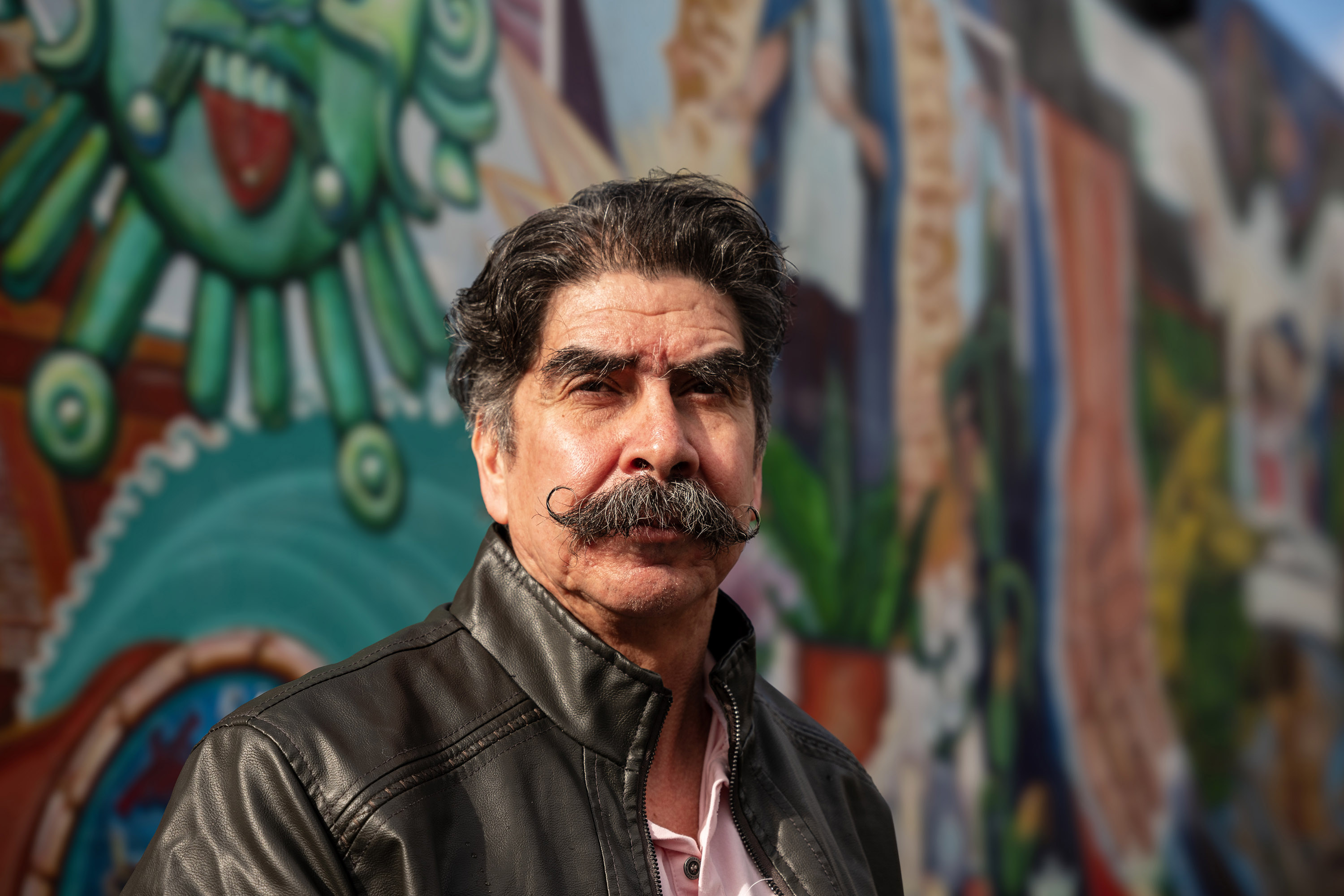 A portrait of Antonio Abundis. He stands in front of a colorful mural on a sunny day.
