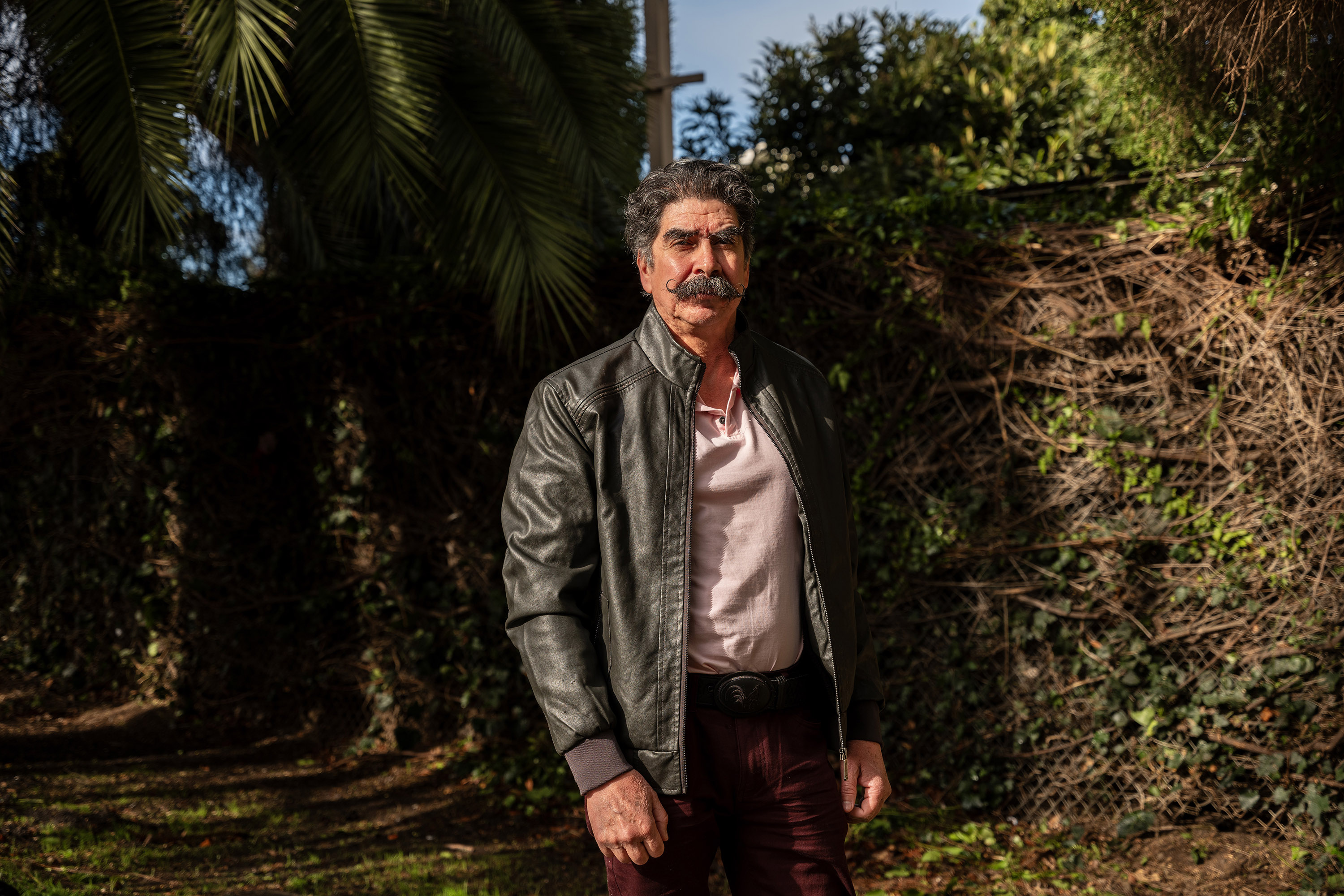 A portrait of Antonio Abundis. He stands outside a lush, green area on a sunny day. Palm fronds and ivy grow in the background.