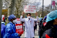Public health workers, doctors, and nurses protest outside a New York City hospital, holding signs. A doctor, wearing a face mask and white coat, is photographed in the center of a crowd. He holds a sign that reads, "If we works ick, you get sick. #PPENOW"
