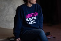 A woman is photographed from the chin down, with the focus on her sweatshirt. The shirt reads, "ABORTION IS ON THE BALLOT."