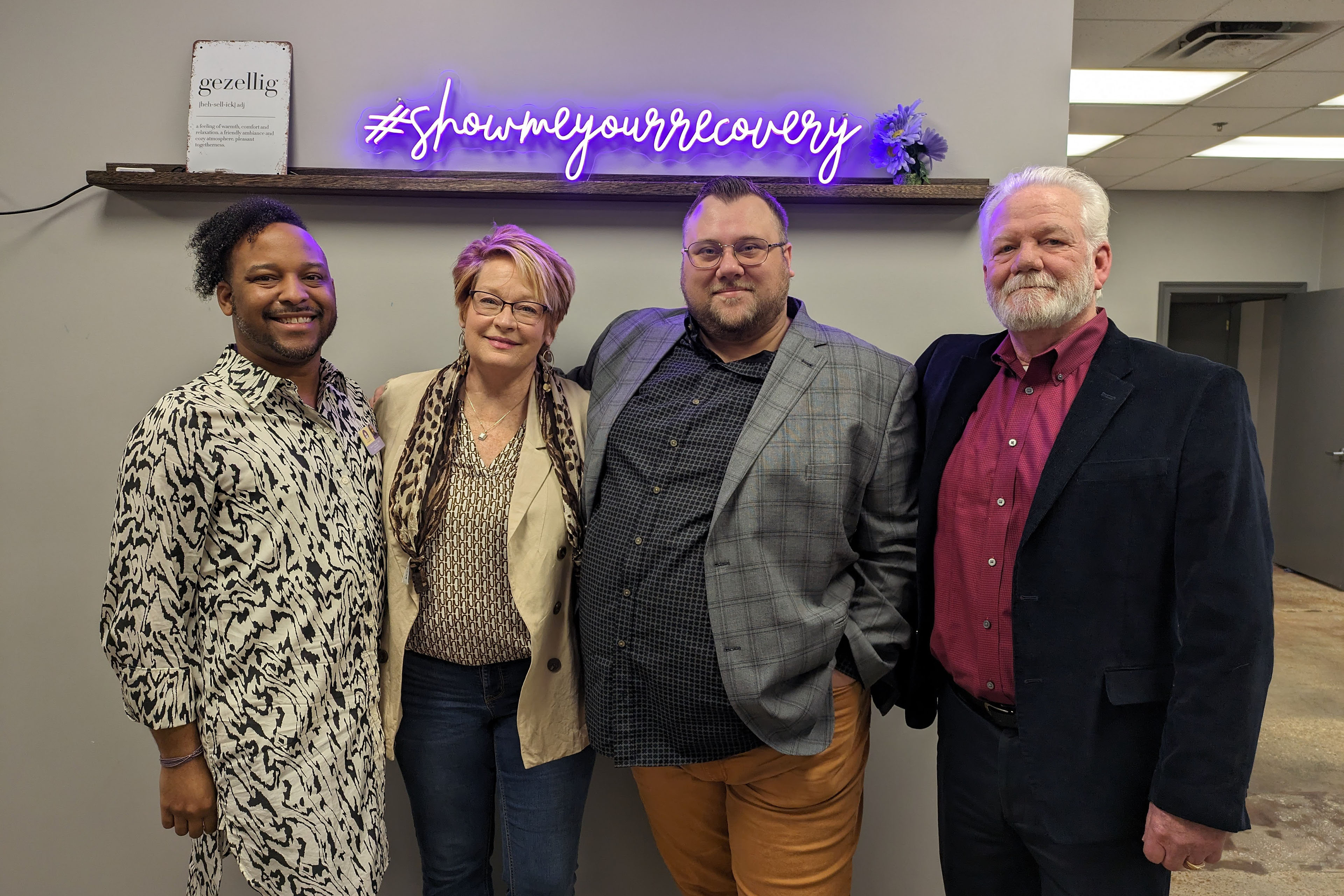 (From left): Devin Burton, Lisa Teggart, Chance Shaw, and Larry Vahle stand side by side in an office room. Above them in a purple neon sign which reads, "#showmeyourrecovery" in script.