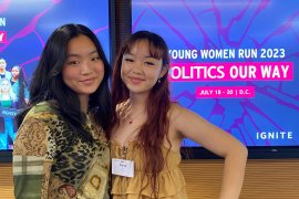 A photo of two teenagers posing for a photo together. They stand in front of a screen that reads, "Women run 2023. Politics our way."