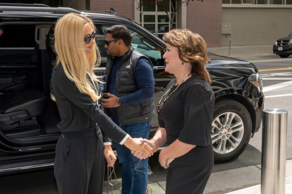 A woman with long straight blonde hair and wearing sunglasses shakes the hand of a woman with shorter brown hair. They are standing outside and beside a black parked SUV.