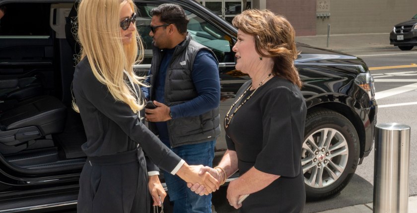 A woman with long straight blonde hair and wearing sunglasses shakes the hand of a woman with shorter brown hair. They are standing outside and beside a black parked SUV.