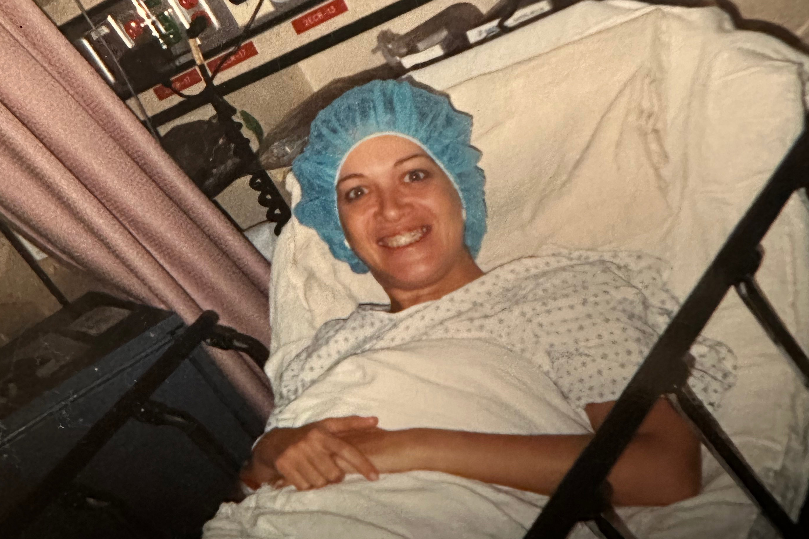 A photo of a woman smiling in a hospital bed with a gauze cap on her head.
