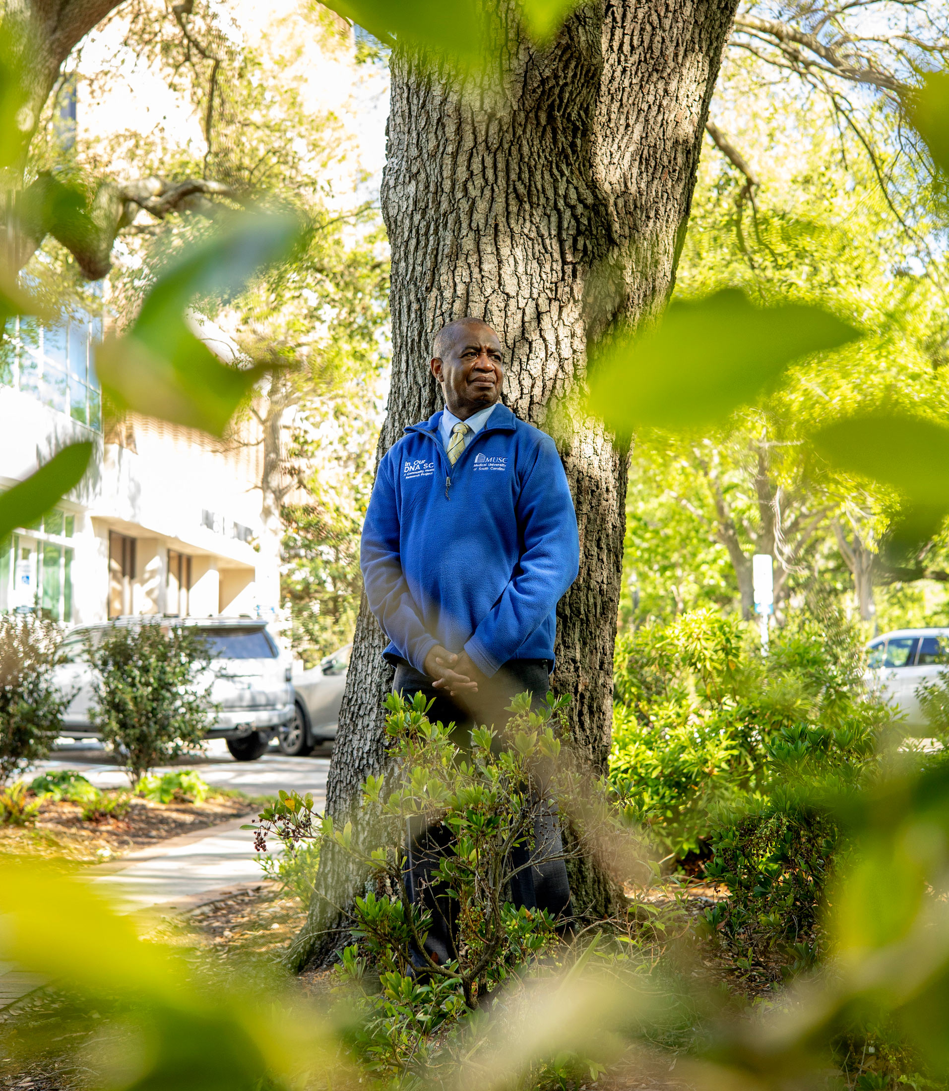 A photo of a Black man in a blue pullover leaning against a tree. Blurred foliage in the foreground frames him.