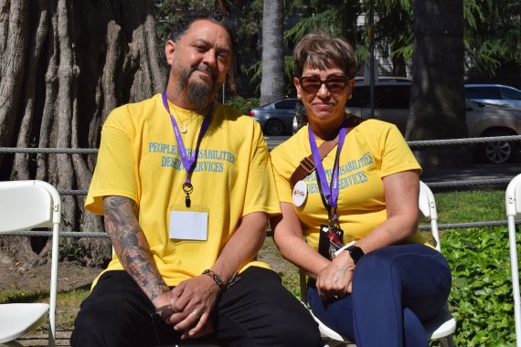 A photo of a man and a woman sitting next to each other outside. They both are wearing yellow shirts that read, "People with disabilities deserve services."