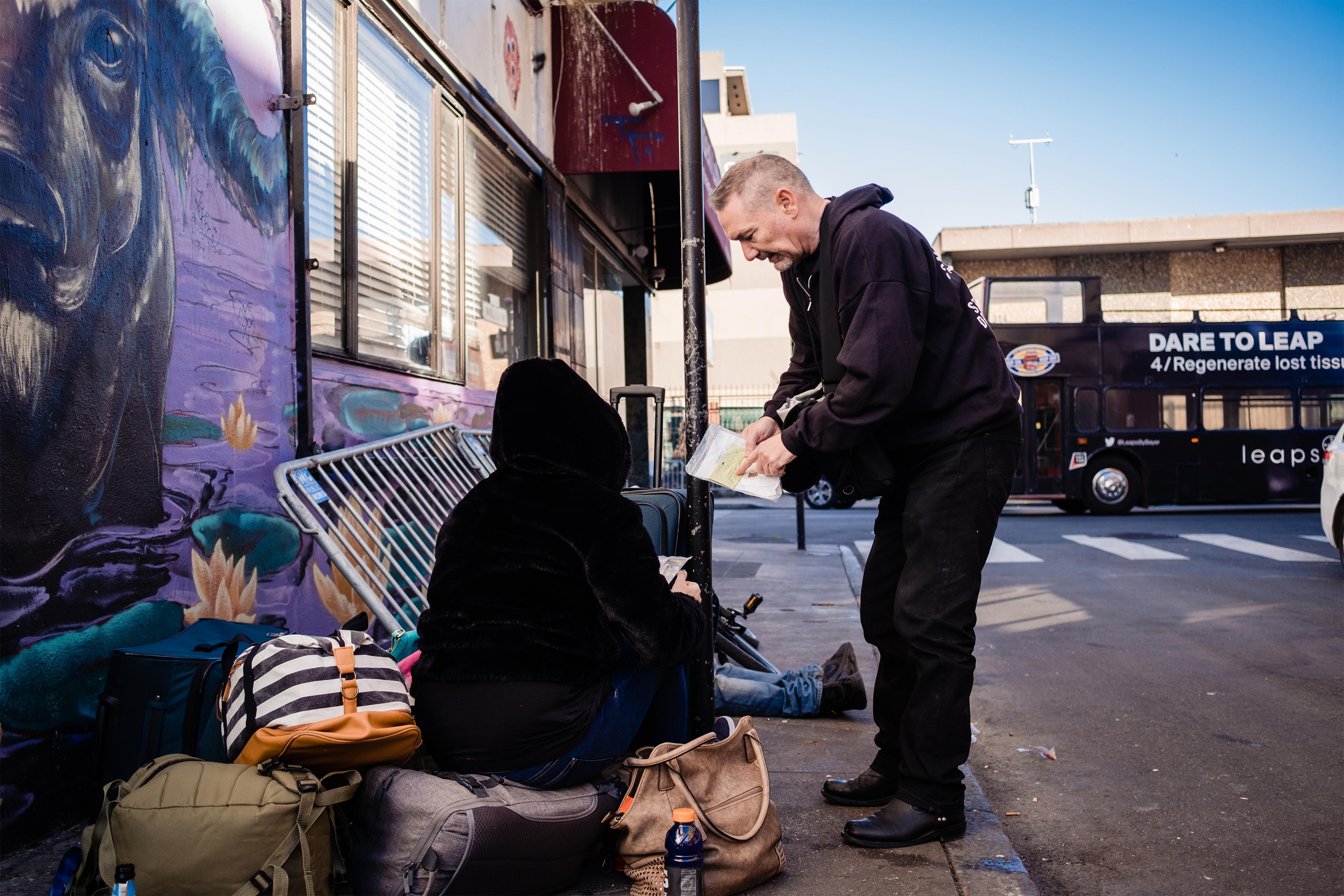 A photo of a man handing out materials to a group of people sitting on the sidewalk.