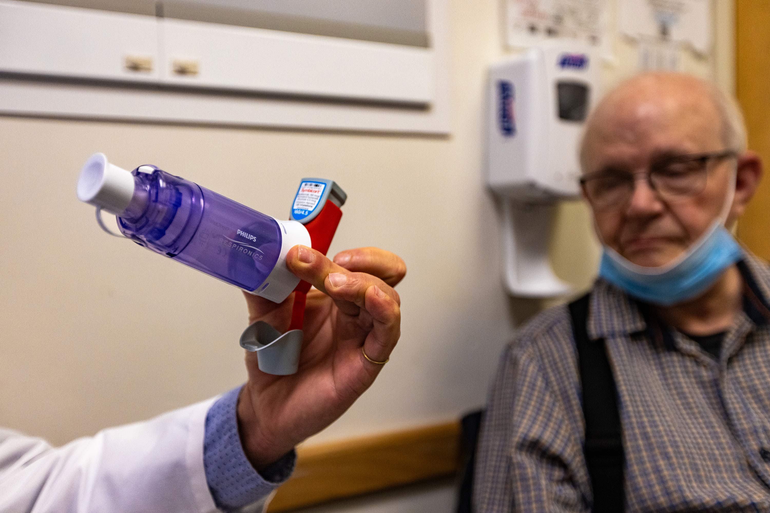 A photo of a doctor's hand holding up a metered-dose inhaler. His patient is seen looking at it in the background.