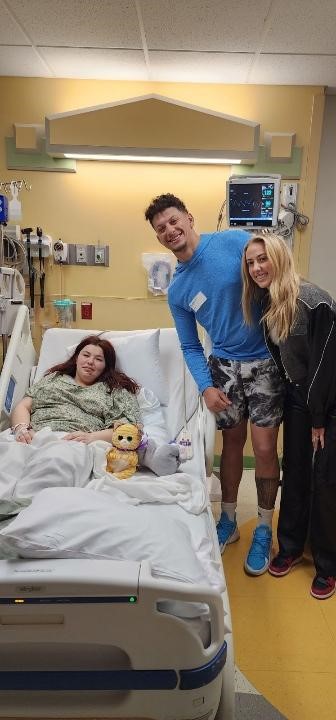 A photo of a woman in a hospital bed. A man and a woman stand next to her, smiling.