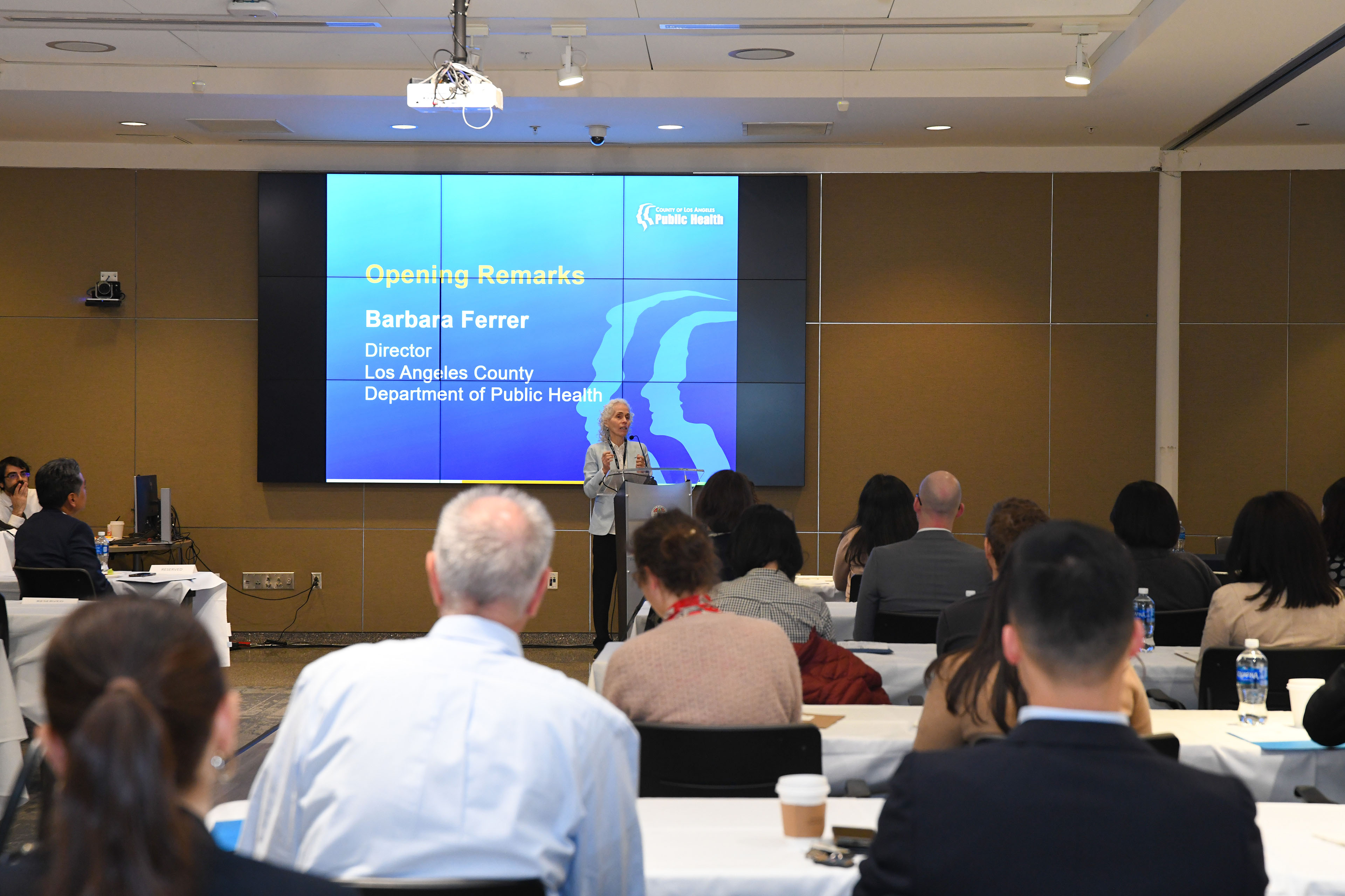 Barbara Ferrer is speaking at a podium at the front of a conference-like room. There is a projector screen behind her. It shows a slide with a blue background and white text that reads, "Opening Remarks / Barbara Ferrer / Director / Los Angeles County Department of Public Health."
