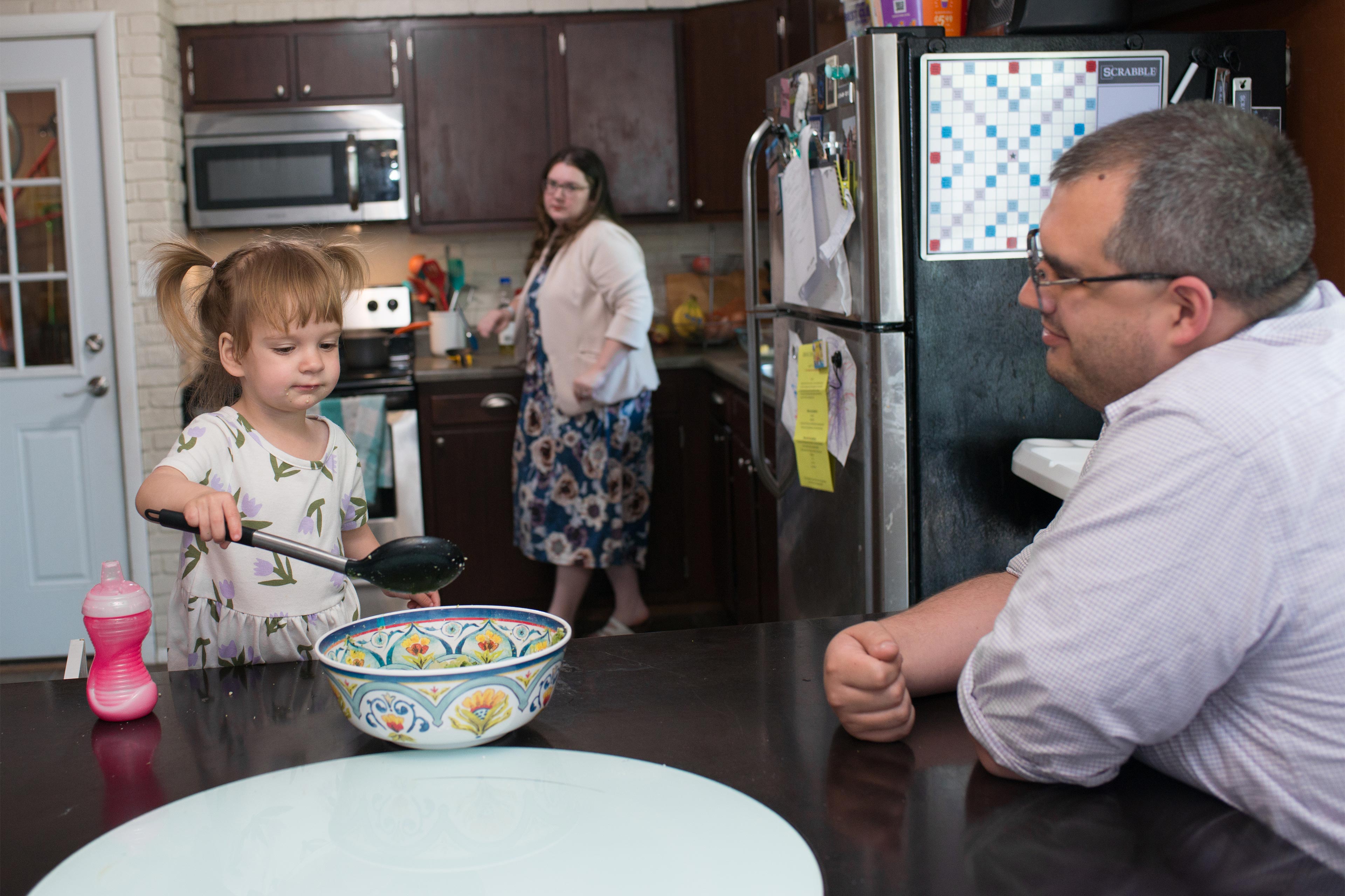 A photo of a young girl with a large spoon and bowl sitting next to her father. Her mother is in the kitchen behind them.